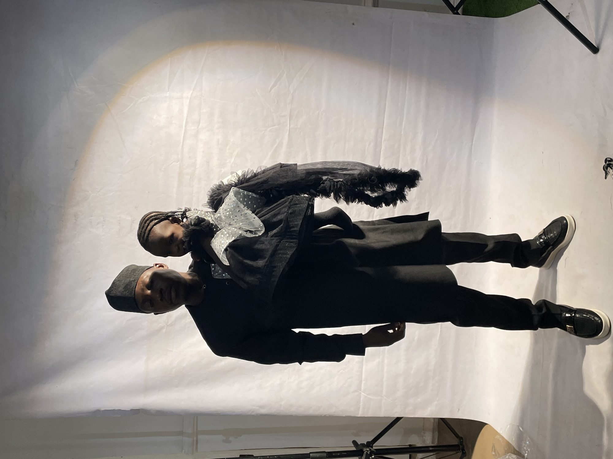 A medium, tilted shot shows a man and child against a white backdrop in what appears to be a studio setting. The man, wearing a black hat and matching attire, carries the child. The child is draped in what looks like a black garment with white polka dots and feather embellishments. Both are facing forward. The environment includes lighting equipment, a white backdrop, and potentially a grassy area visible in the upper right corner. The location is reported as Barakin Akawo, Nigeria.