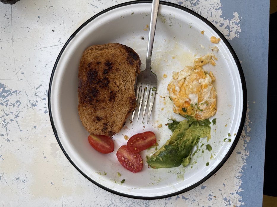 A white enamel plate holds a partially eaten breakfast. A slice of browned toast sits on the left, next to three halves of cherry tomatoes. A silver fork rests between the toast and a portion of scrambled eggs with green herbs. Sliced avocado is arranged near the eggs. The plate shows signs of food residue and sits on a textured surface with a blue and white pattern.