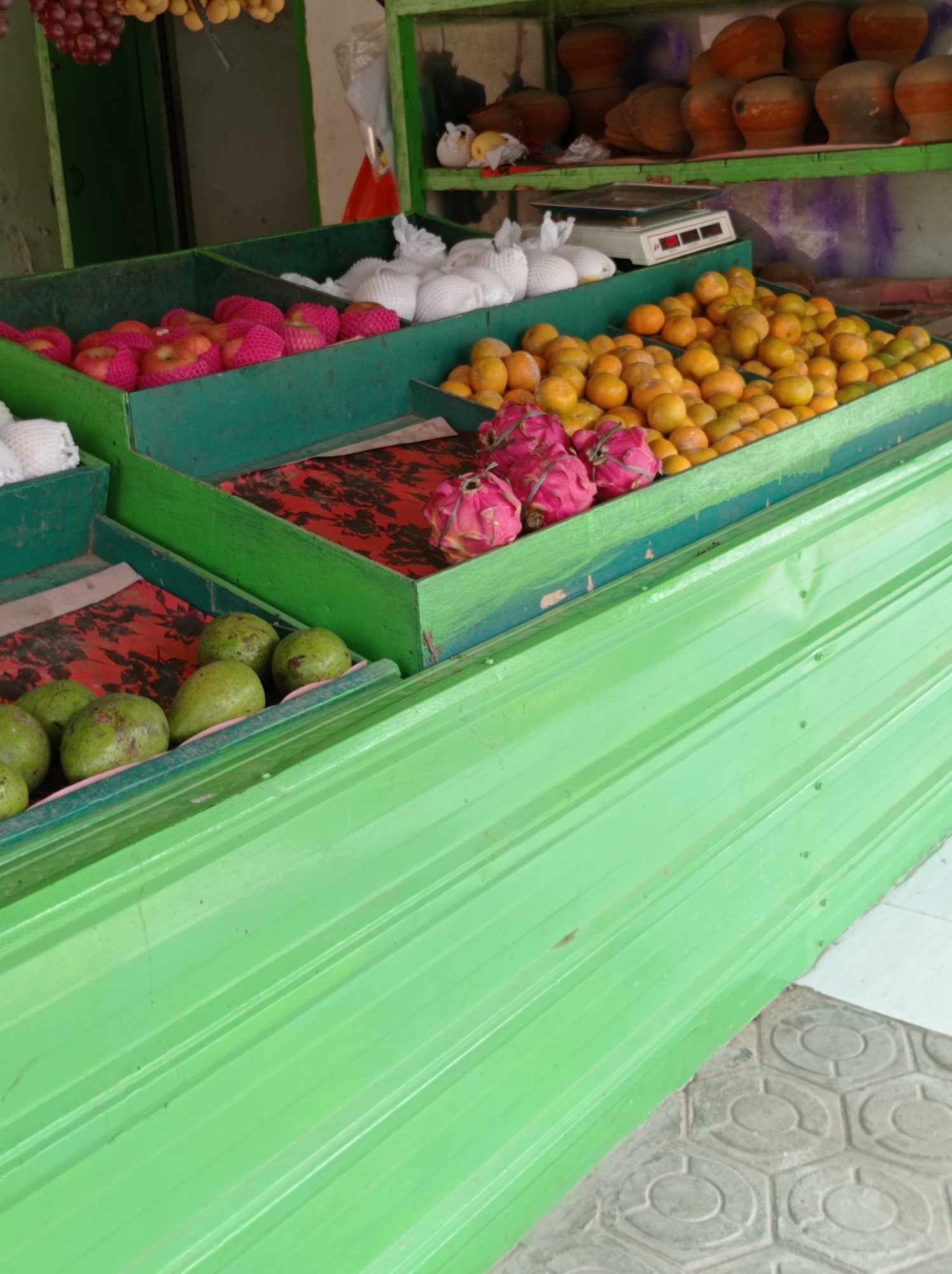 An outdoor fruit stand in Kramat, Indonesia, displays a variety of fruits in green wooden crates. In the foreground, a row of green avocados rests on a red floral patterned cloth. Behind them, in a higher crate, several pink dragon fruits are visible. Further back, a large quantity of small, bright orange citrus fruits, likely tangerines or mandarins, fill another crate. Above the citrus, a white digital scale is positioned next to a display of white-wrapped fruits, possibly pears or apples. In the uppermost crates, a collection of red apples, also wrapped in pink netting, are arranged, alongside more white-wrapped fruit. The background reveals a shelf with terracotta pots and other items, as well as a green painted wall. The stand is constructed with green corrugated metal sheeting.