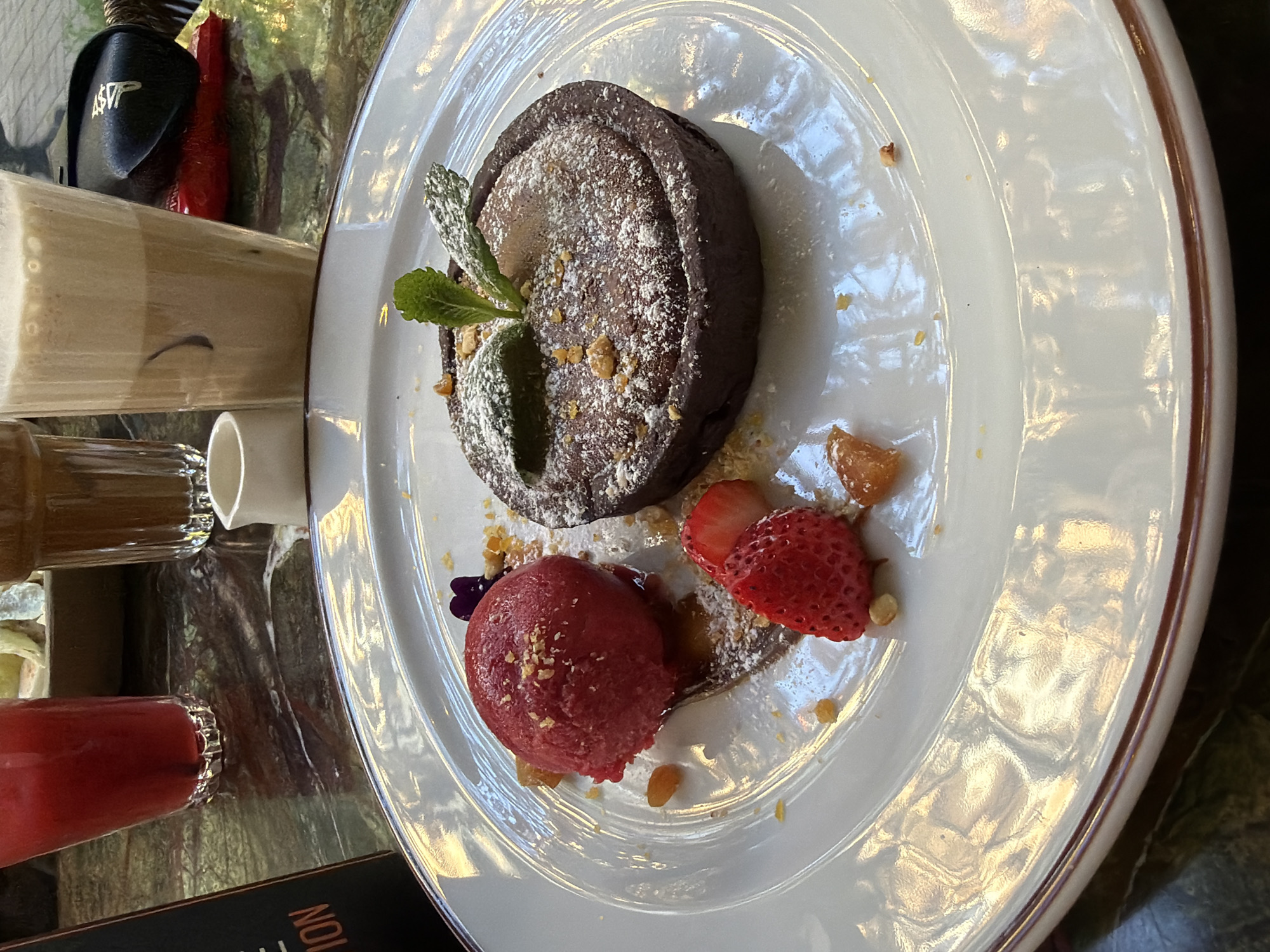 A chocolate dessert is presented on a white plate with a brown rim. The dessert features a round, dark chocolate cake dusted with powdered sugar and garnished with a sprig of mint. Next to the cake is a scoop of red sorbet and sliced strawberries. The plate rests on a reflective surface that appears to be a marble or polished stone tabletop. To the left are glasses, one containing a light-colored beverage and another with a red liquid. The scene gives the impression of being in a restaurant or cafe. This is likely in the city of Sofia, Bulgaria, as requested.