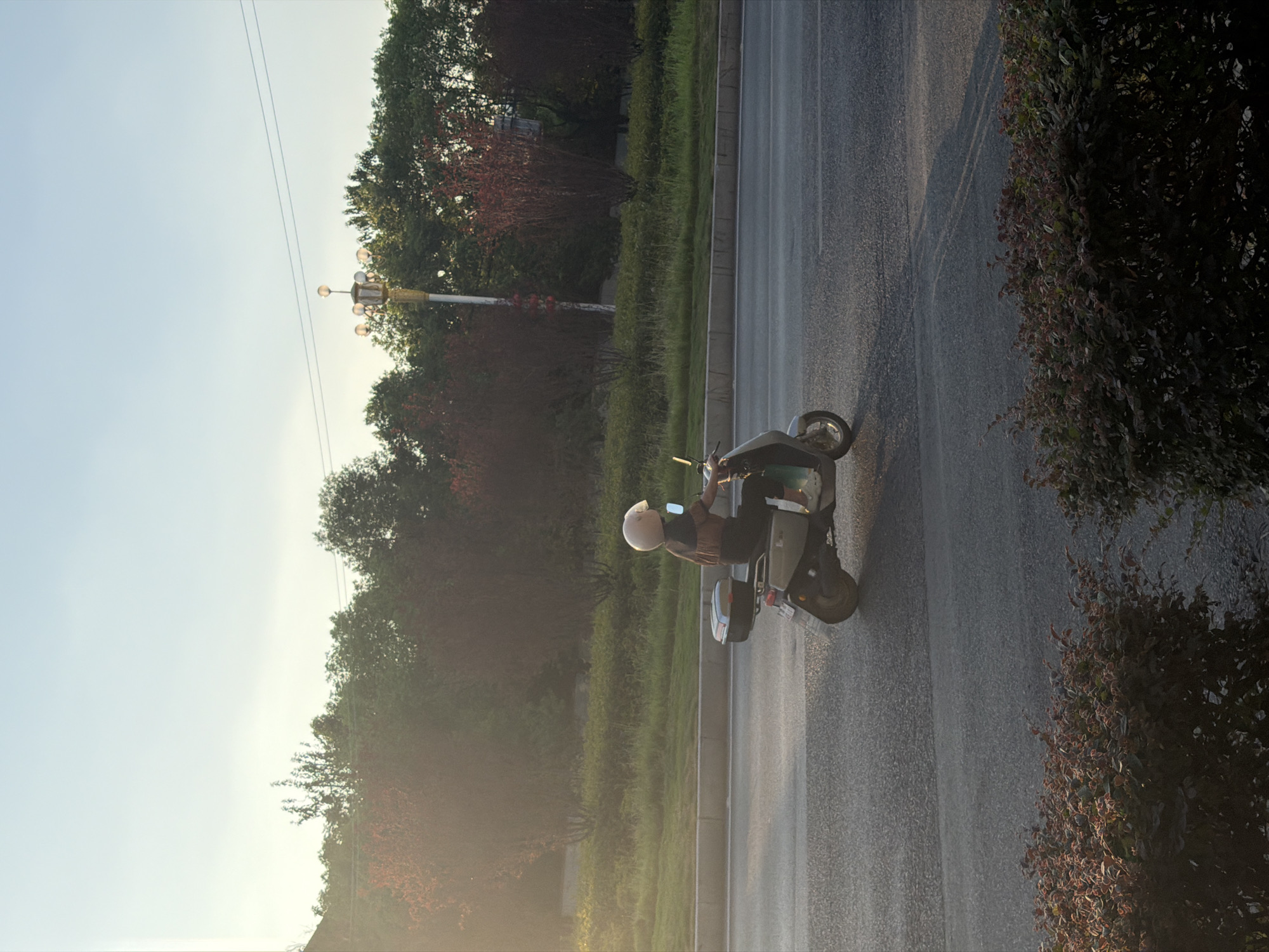 A person is riding a scooter on a road in Jiangjin, China, in the late afternoon. The sun is low in the sky, casting long shadows. The scooter is light-colored, and the rider is wearing a helmet and a striped shirt. The road is paved with asphalt, and there is a sidewalk to the left. Behind the sidewalk, there is a row of trees and bushes. In the background, there is a street lamp with multiple lights. The sky is a light blue with some clouds. The overall scene is peaceful and serene.