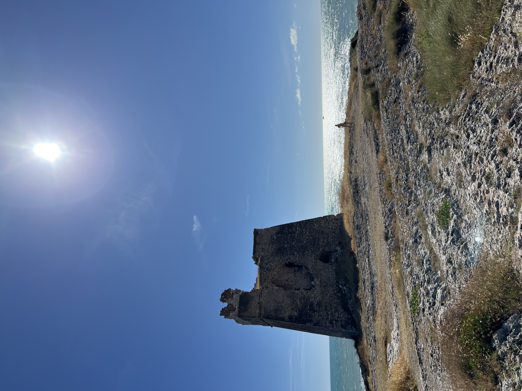 A wide, low-angle shot captures the ruins of an ancient stone tower on a rocky cliff overlooking the sparkling Adriatic Sea under a bright, cloudless blue sky with the sun prominently featured. The tower, made of weathered stone, stands on the left side of the frame, displaying significant damage with crumbled sections at its top and openings in its walls. The rocky terrain leading up to the tower is sparsely covered with dry grass and some small green plants. In the distance, a lone figure stands facing the sea, their back to the camera, appearing to be a person wearing a swimsuit. The sun's glare creates a bright, shimmering effect on the surface of the water. The overall scene evokes a sense of solitude and the passage of time, typical of coastal historical sites near Peschici, Italy.
