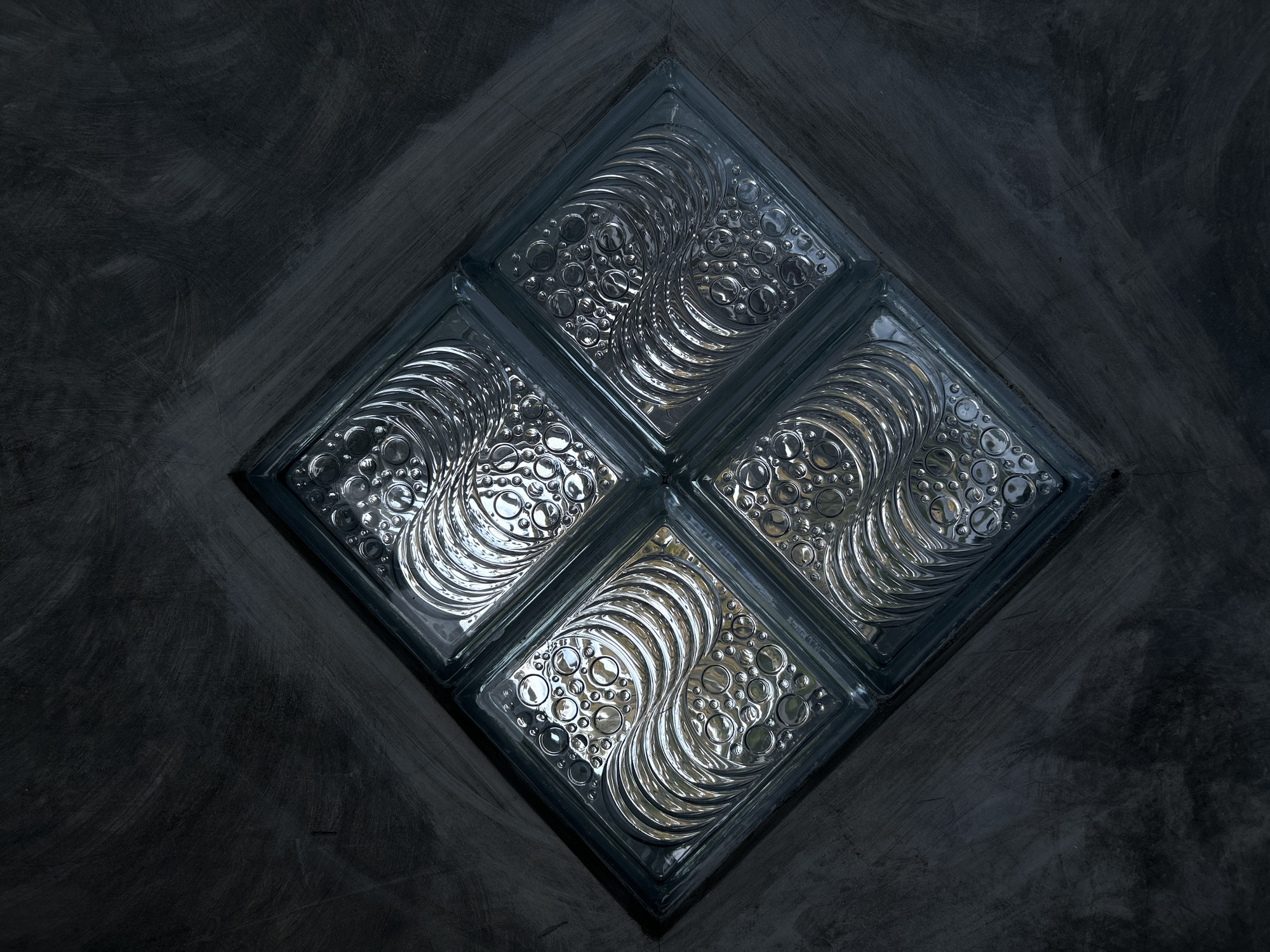 The image depicts a square formation of four glass blocks with a swirl and bubble pattern embedded within. The glass blocks are set into a dark, textured, possibly concrete or plaster wall, arranged in a diamond orientation. There are no visible people in the scene. The location of Lhokseumawe, Indonesia is not evidenced in the image.