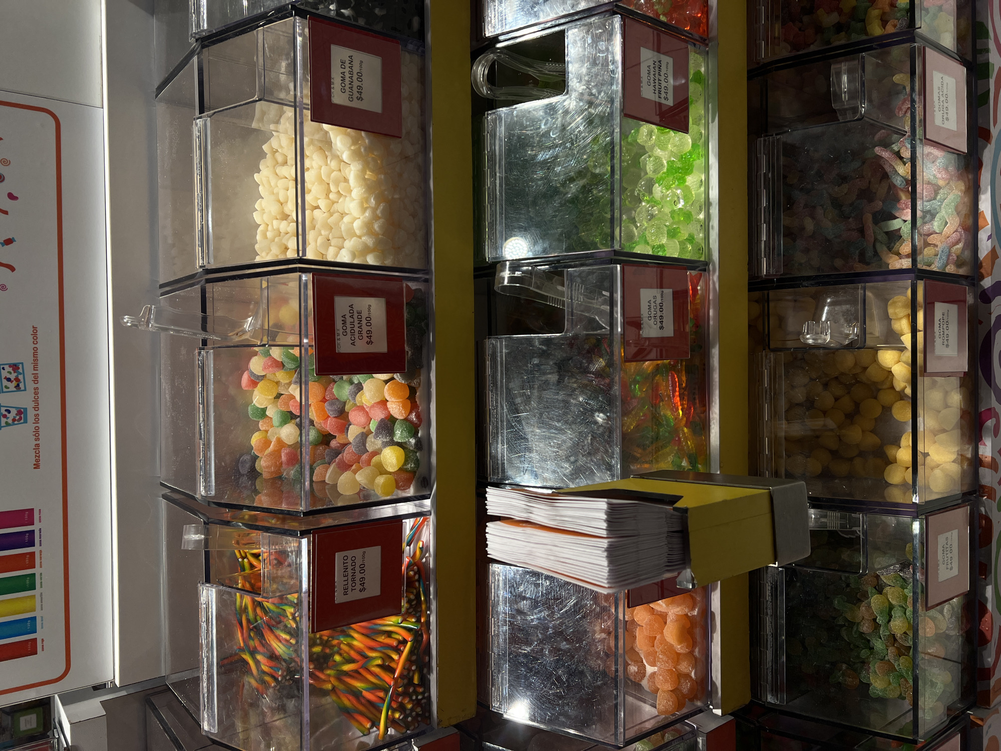 A medium shot shows several candy dispensers at a store in Mexico City, Mexico. The dispensers are clear, square-shaped containers with black trim, and they are filled with various types of colorful gummy candies, including gummy worms, gummy bears, and other assorted shapes. Each dispenser has a label indicating the type of candy and its price per 100 grams, which is $49.00.
On the left side of the frame is a colorful poster with Spanish text, "Mezcla sólo los dulces del mismo color", which translates to "Mix only the candies of the same color". A stack of white paper napkins sits on a dispenser dividing the gummy candies. The overall lighting is bright, and the colors of the candies are vibrant and appealing. The scene suggests a candy store or section within a larger retail establishment.