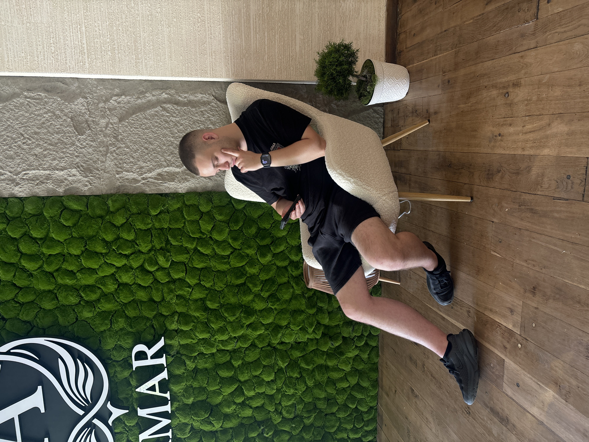In Tsarevo, Bulgaria, a young man with a shaved head and a black t-shirt and shorts is seated in a cream-colored armchair. He holds a smartphone in his left hand and rests his right hand on his chin, appearing to be looking at the phone. The armchair is situated on a wooden floor in front of a wall covered in vibrant green moss. To the right of the armchair, a small bonsai tree in a white pot is visible. Behind the moss wall, a section of rough grey stone and a textured cream panel are present. The scene suggests an indoor setting, possibly a lounge or reception area.
