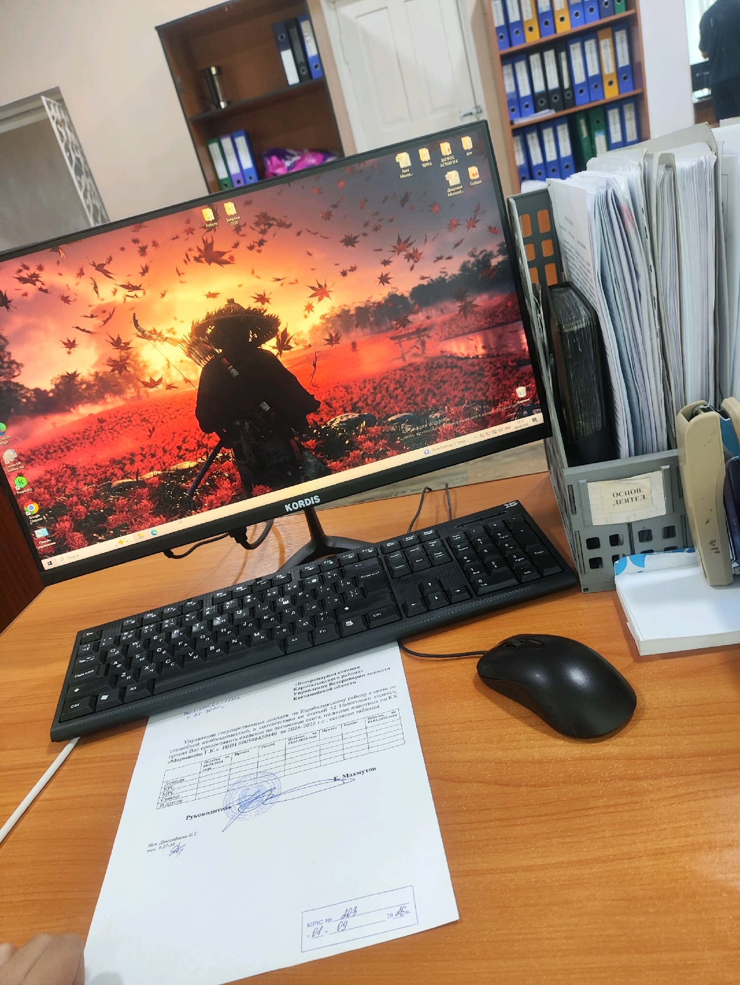 An indoor, medium shot captures a desk with a computer monitor displaying a red and orange landscape with a silhouetted figure. The monitor is a KORDIS brand. To the right of the monitor, a stack of files and documents is organized in a gray plastic holder. A black keyboard and mouse are positioned in front of the monitor. Below the keyboard, a sheet of paper with text and a stamp is laid out. The desk surface is made of wood. In the background, a shelf unit is filled with various colored binders and folders, with one binder in the foreground having a label that reads "ОСНОВ. ДЕЯТЕЛЬ.". The environment appears to be an office setting. The text on the paper suggests a location in Qarabalıq, Kazakhstan, with mentions of "Карабалыкскому району" (Qarabalıq district) and "Костанайской области" (Kostanay region).