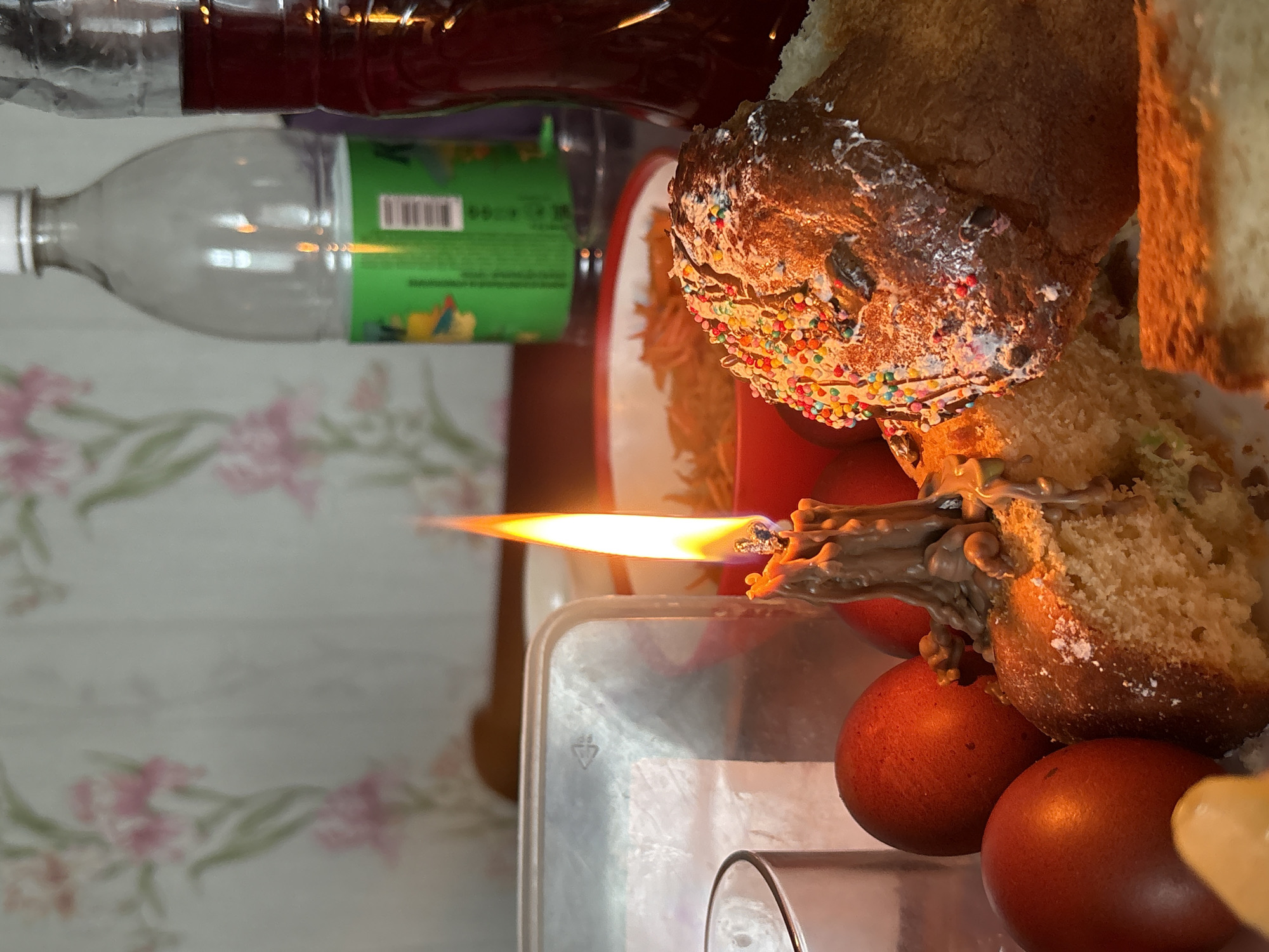 The image depicts a celebratory scene, possibly Easter. A partially sliced and decorated sweet bread (Kulich) sits atop a plate, alongside dyed red eggs. A lit candle is placed among the Kulich and eggs. Glass bottles are present in the background, along with a floral patterned wall. The location, Пожарское сельское поселение, Ukraine, is provided.