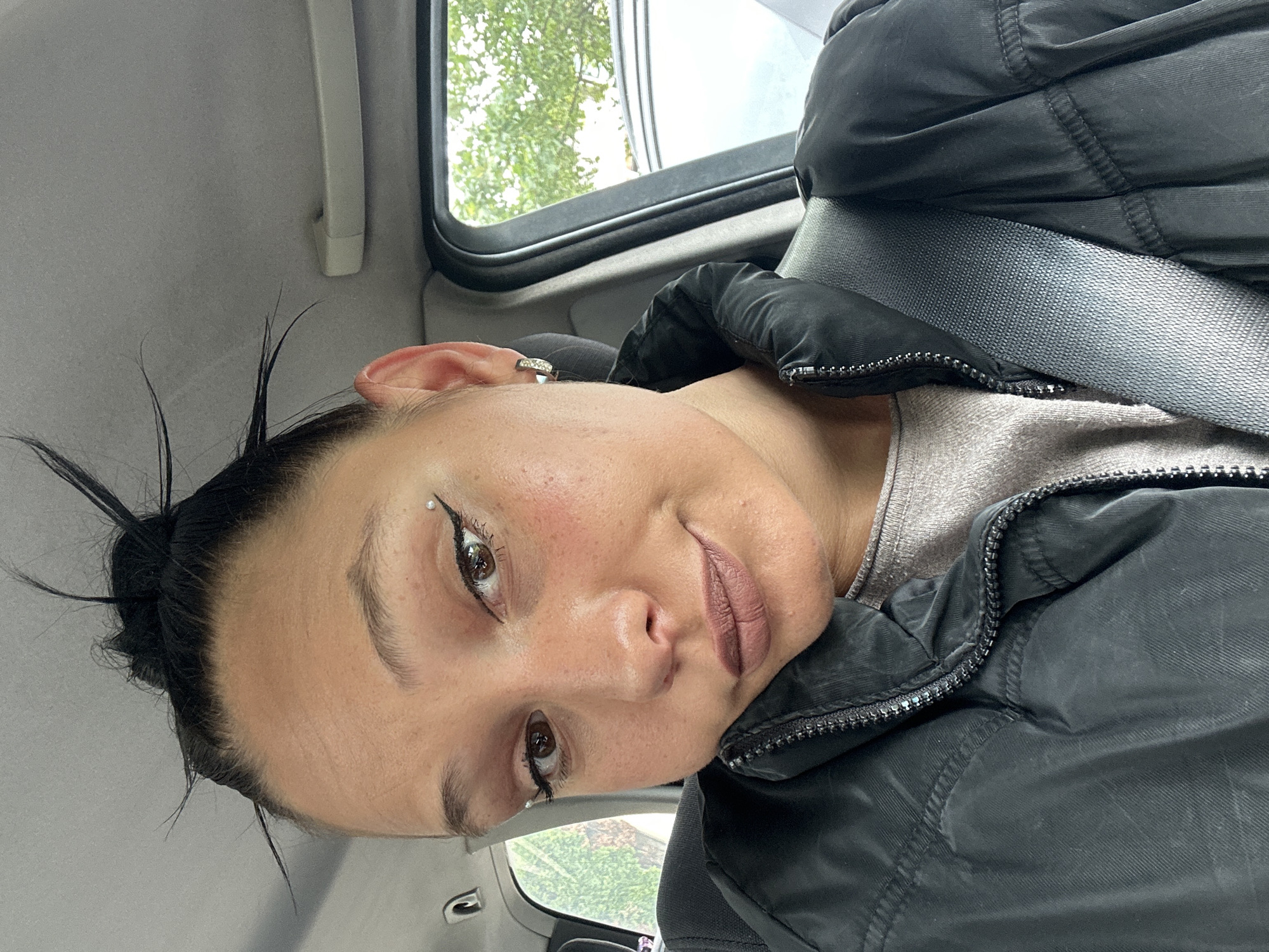 A woman is in a car, wearing a black puffer jacket and a seatbelt. She has dark hair pulled back, and her skin tone is fair. She has a piercing above her left eyebrow and is wearing light lipstick. The background shows a window with green trees and a gray sky. The car's interior is visible, including the headliner and door frame. The image does not provide any information to determine the location in Sofia, Bulgaria.