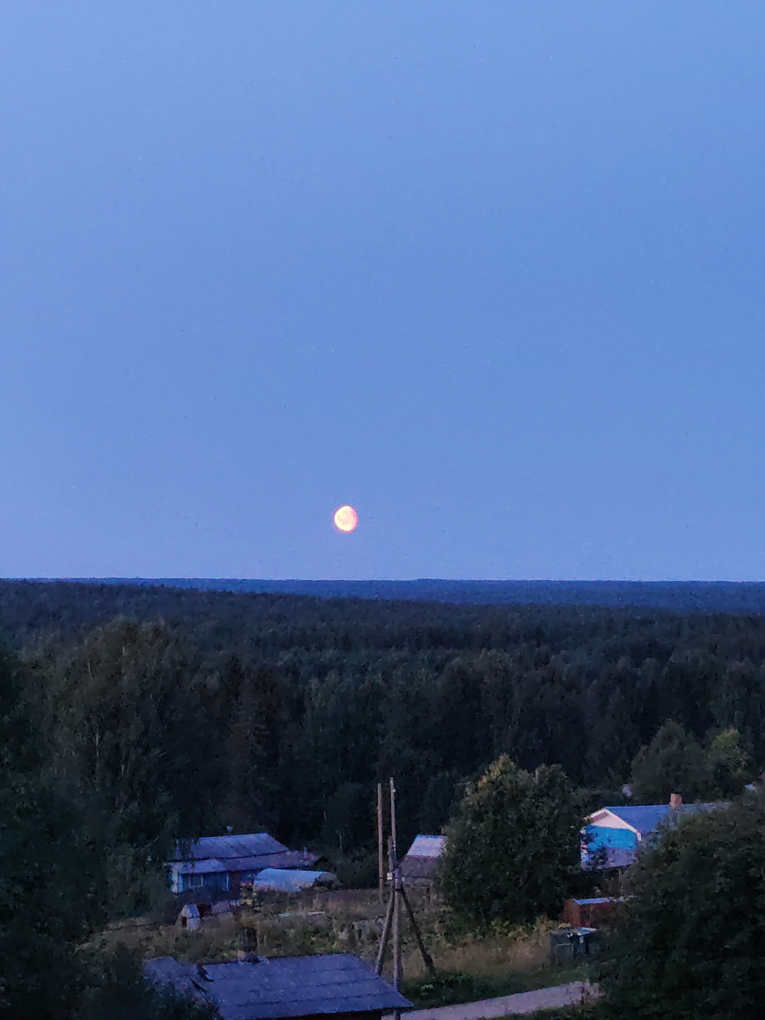 The image is a vertical shot taken at dusk or dawn, depicting a rural landscape under a clear, pale blue sky. A glowing, orange-red moon is visible high in the sky, slightly to the left of center. Below the sky, a dark, dense forest stretches across the horizon. In the foreground, nestled amongst vegetation and trees, are several small, weathered houses with blue roofs. The structures appear to be simple, rustic dwellings. A wooden utility pole with wires is positioned to the right of the houses. The overall impression is one of a quiet, isolated, and somewhat melancholic rural scene in the city of Chukhlem, Russia.