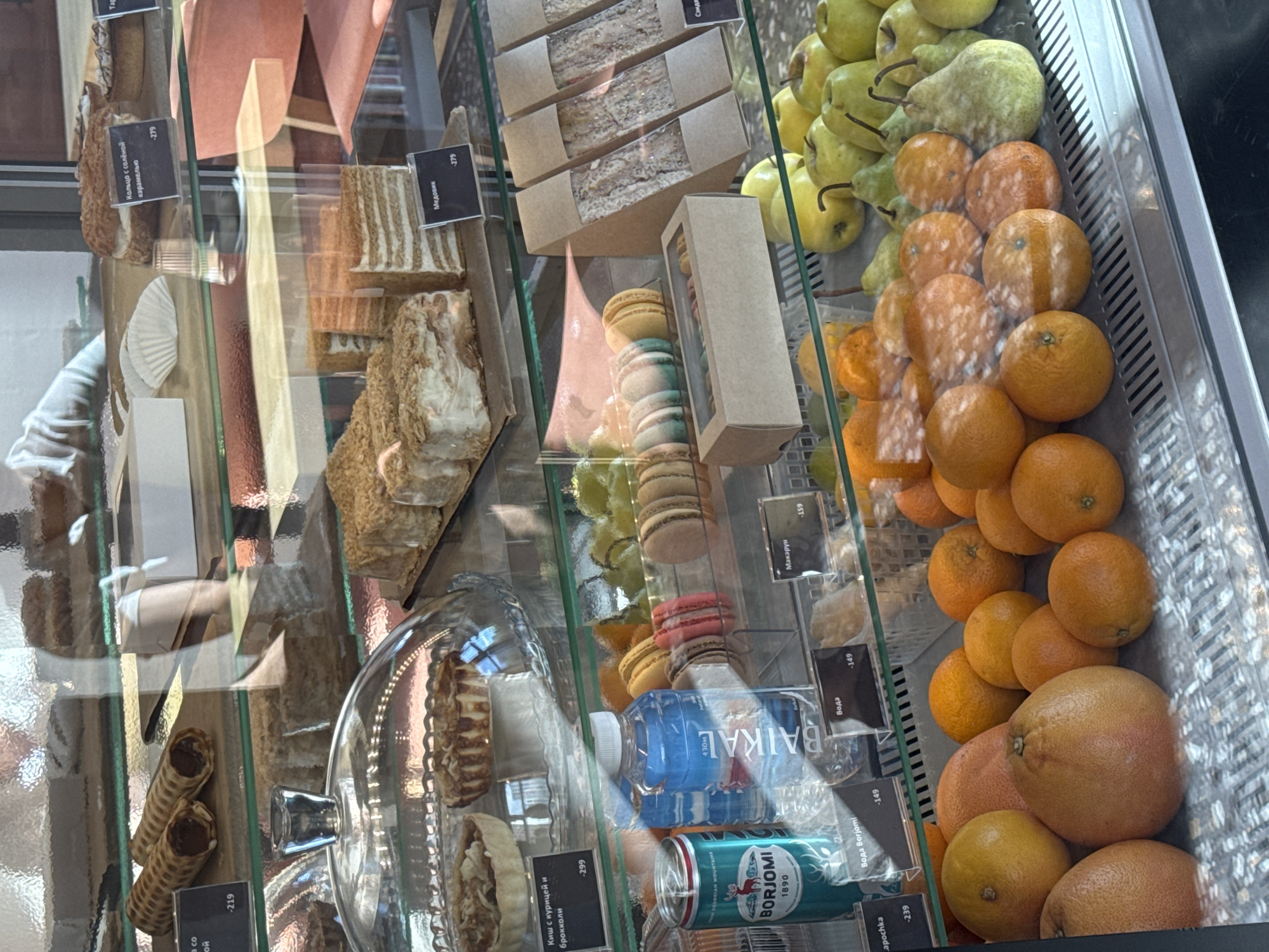 The image shows a display case filled with various food items, including pastries, sandwiches, macarons, fruits, and bottled drinks, likely in a bakery or cafe. A glass partition separates the viewer from the merchandise. There are price tags visible in front of some items. The lighting is artificial, creating reflections on the glass. In the background, blurred figures and furniture suggest an indoor commercial setting. The specific location in Patrusheva, Russia, is not discernible from the image itself, but the presence of Russian language on some labels confirms a Russian-speaking context.