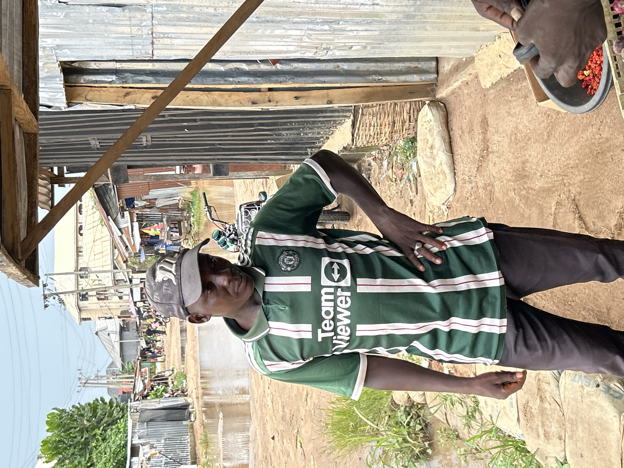 An outdoor, brightly lit scene in what appears to be a market or informal settlement in Ihiagwa, Nigeria. The main subject is a man standing in the foreground, facing slightly towards the camera with a neutral expression. He wears a dark green and white striped polo shirt, which features the "Team Viewer" sponsor logo and what appears to be a football club crest. He also wears a gray cap with an indiscernible logo and dark trousers. His right hand rests on his stomach, while his left arm hangs loosely.
To the right of the man, a dark-skinned hand is visible, holding a black mortar and pestle or a similar grinding tool. Inside the tool are numerous bright red chili peppers or spices, suggesting a food preparation or vending activity. In the immediate foreground, a beige sack, possibly cement, lies on the ground, with a partially visible word that might be "CEMENT". The ground is mostly dry, light brown earth, uneven and somewhat dusty, with patches of green grass and small puddles of water in some areas.
The background reveals a bustling environment with structures made primarily of corrugated metal sheets and wooden planks, indicative of informal stalls or simple buildings. Power lines and utility poles are visible against a bright, clear sky in the distance. A motorbike or moped is parked further back on the left. In the far distance, more buildings and blurry figures of people suggest a lively street or market atmosphere. The strong sunlight and clear sky indicate a daytime setting, likely mid-morning or early afternoon.