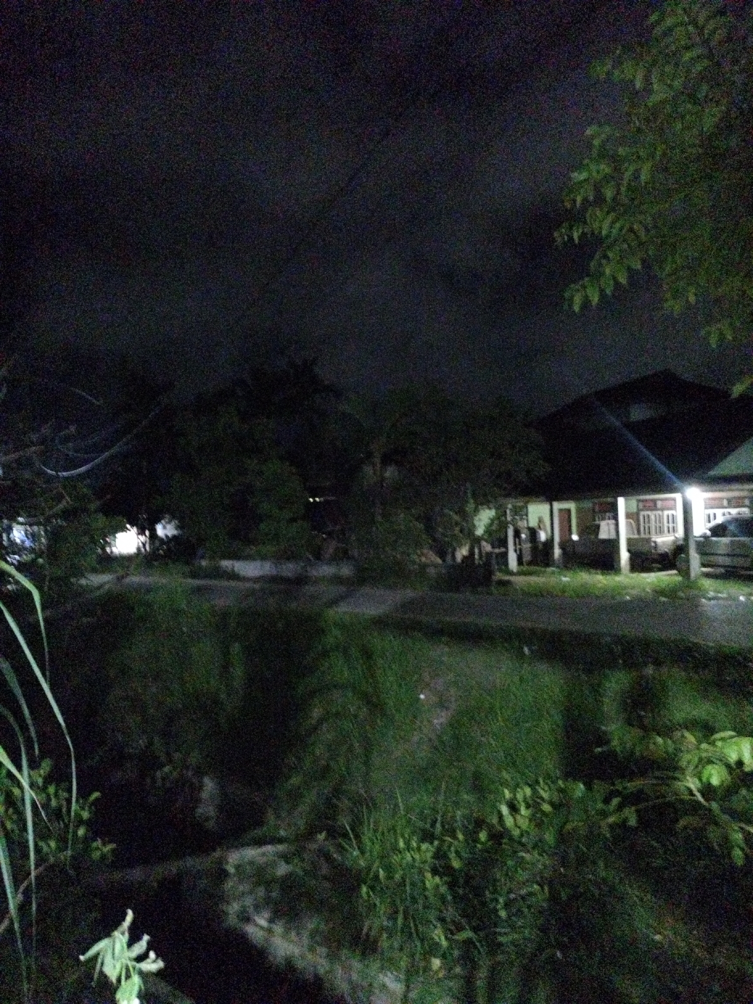 The image depicts an outdoor night scene in Seuneuboklhong, Indonesia. A one-story house with a covered porch is visible in the background, illuminated by interior lights. People appear to be standing or sitting on the porch. A vehicle is parked adjacent to the house. In the foreground, there is dense vegetation including tall grasses and various plants. A road or walkway is situated between the vegetation and the house. The sky is dark with cloud cover. Electrical wires are visible strung across the sky. The image is grainy, suggesting low-light conditions.
