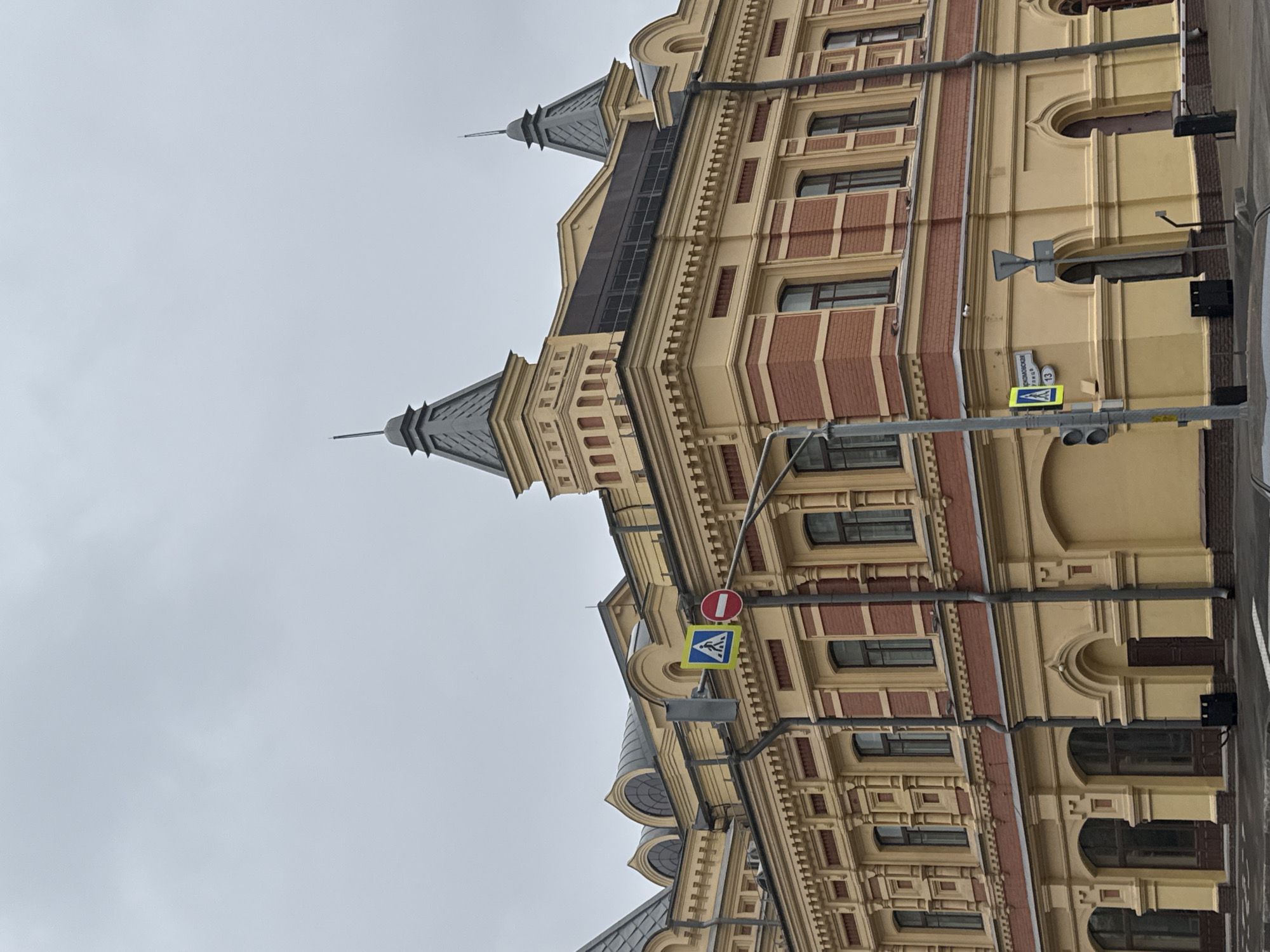 The image shows a street view of a building in Nizhny Novgorod, Russia. The building is a multi-story structure with a yellow facade and red brick accents. It features ornate architectural details, including columns, cornices, and arched windows. The roof has several conical towers with spires. In the foreground, there are traffic lights and street signs, one of which indicates "Комсомольская улица 13" (Komsomolskaya Street 13). The sky is overcast, suggesting cloudy weather. There are no people visible in the image. The scene appears to be a typical urban streetscape.