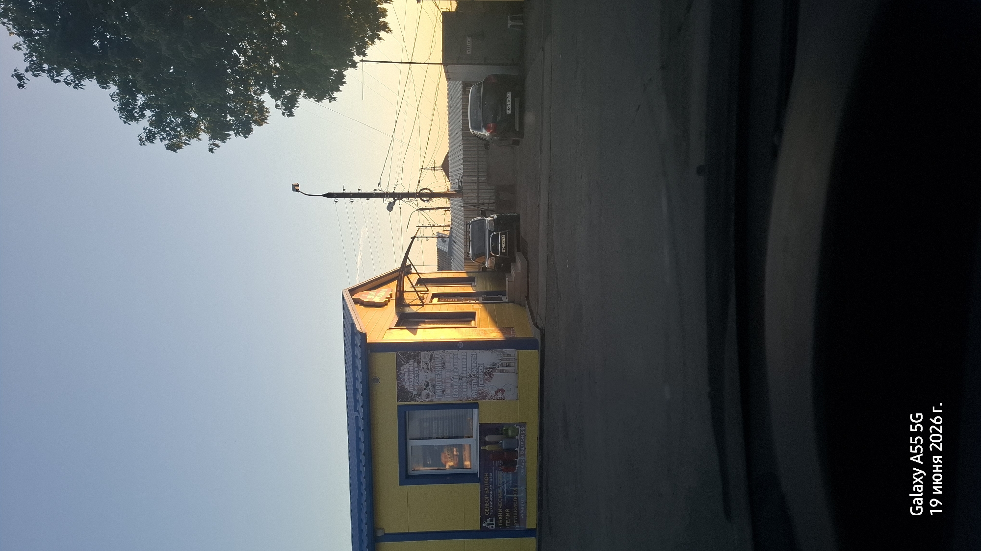 The image depicts a small yellow commercial building with a blue roof, likely a shop, situated in an outdoor area. The building has a large window and an awning over the entrance. Various signs are visible on the building, some of which appear to be in Russian. Two vehicles, a dark SUV and a sedan, are parked in front of the building. In the background, there are utility poles with wires, a metal fence, and a large tree on the right side of the frame. The scene is illuminated by the warm, low-angled light of what appears to be either sunrise or sunset. The foreground shows the interior of a vehicle, suggesting the photo was taken from inside a car. The bottom of the image contains a timestamp indicating "Galaxy A55 5G 19 июня 2026 г.", which translates to "Galaxy A55 5G June 19, 2026." The location is identified as Rostov-on-Don, Russia.