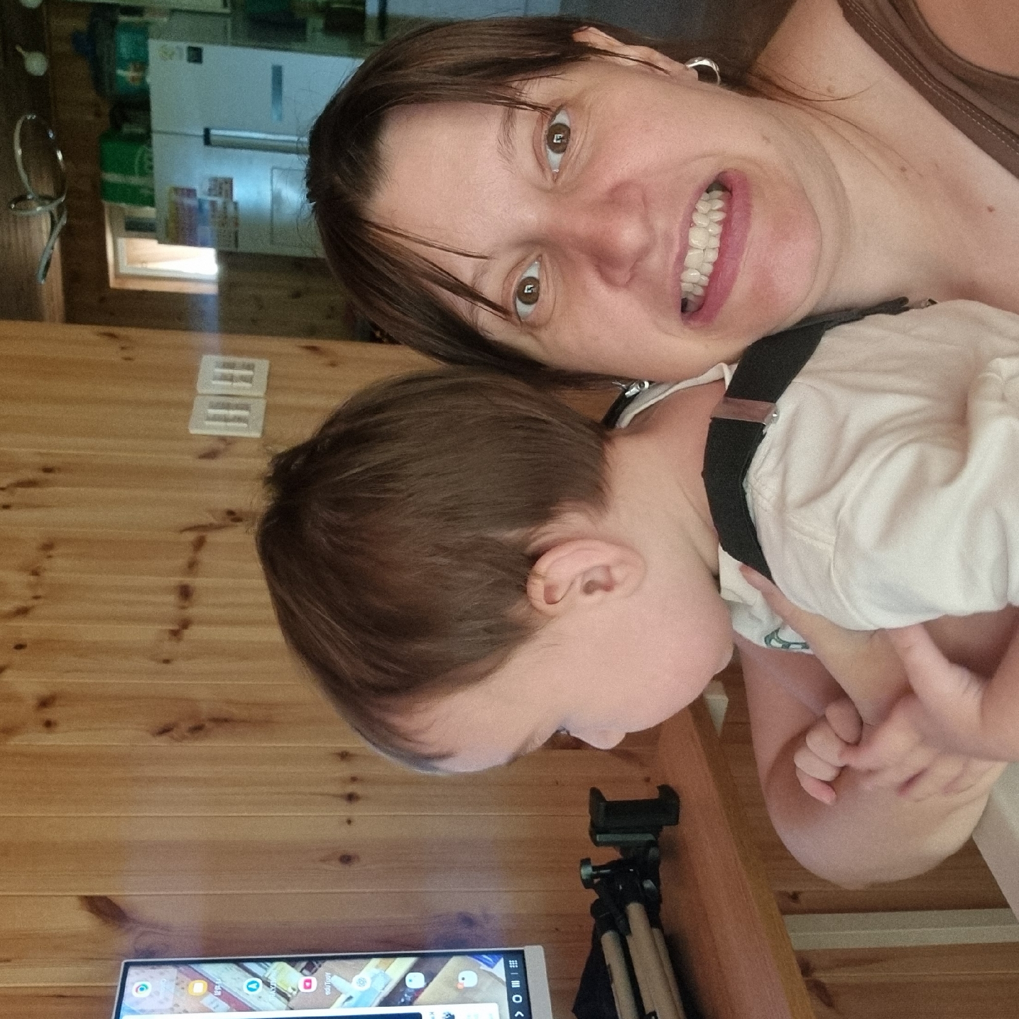 A medium shot captures a woman with brown hair and brown eyes making a contorted facial expression, her mouth open and teeth bared. She is holding a young child in her arms, whose face is turned away from the camera, looking towards a television screen. The television is displaying various icons, suggesting it is a smart TV or tablet. In the background, wooden paneled walls are visible, along with a white refrigerator and kitchen cabinets. A tripod is placed on a wooden surface to the left of the television. The overall scene appears to be in a rustic indoor setting.