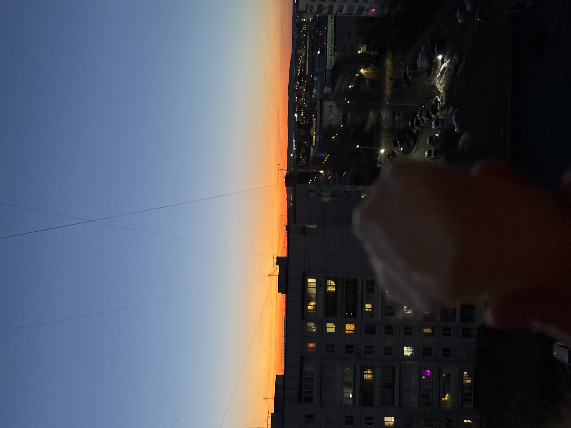 The image shows a nighttime urban scene in what appears to be Chaikovsky, Russia, as evidenced by the architecture and urban layout. The view is from a high vantage point, likely a building, looking out over a city landscape. The sky transitions from a deep blue to a vibrant orange-red, indicating either sunset or sunrise. Power lines stretch across the sky. Buildings are interspersed throughout the scene, with lit windows indicating activity within. In the foreground, a blurry object is visible, possibly being held up to the camera. Beyond the immediate foreground, roads and parking areas are visible, with parked cars illuminated by street lights.