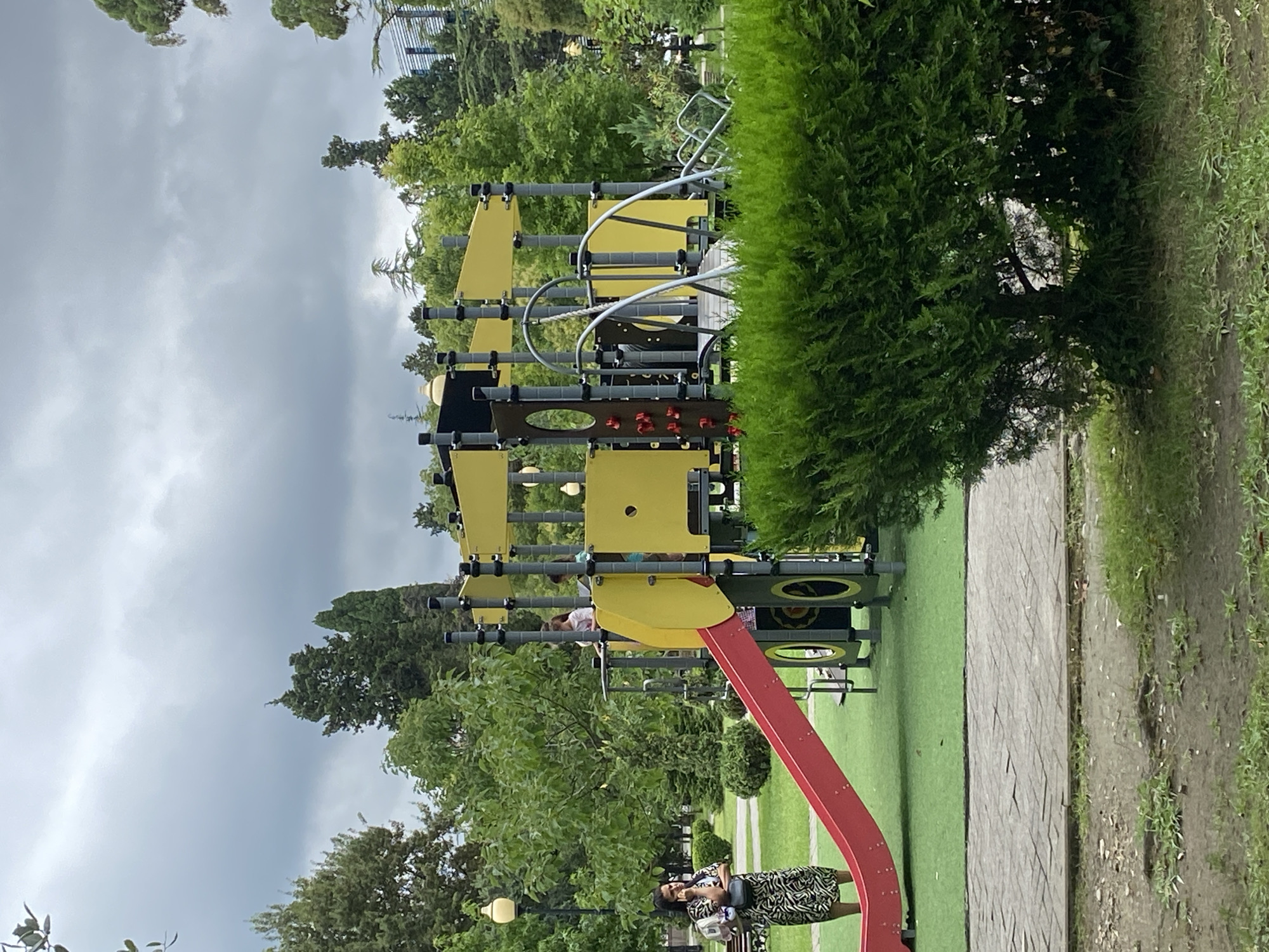 A playground set is situated in a park in Sokhumi, Abkhazia, under a cloudy sky. The playground has yellow and grey structures, a red slide, and various climbing features. A woman in a patterned dress is standing near the slide, looking towards the left, with a backpack on her shoulder. Two children are visible on the playground structure. Lush green trees and bushes surround the playground, and a paved path is visible in the foreground. The ground appears to be a mix of grass and dirt.