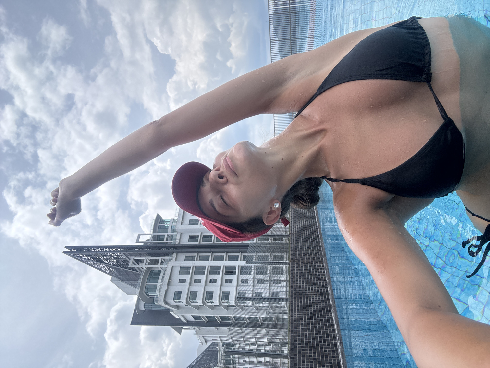 The image depicts a woman in a black bikini and a red cap, seemingly in a rooftop pool in Kuala Lumpur, Malaysia. She is leaning back with her right arm outstretched towards the cloudy sky. Her face is turned upwards, eyes closed. The pool is tiled with blue mosaics, and behind her, a tall, modern building is visible, partly obscured by the water's edge. The overall impression is of a relaxed, urban setting.