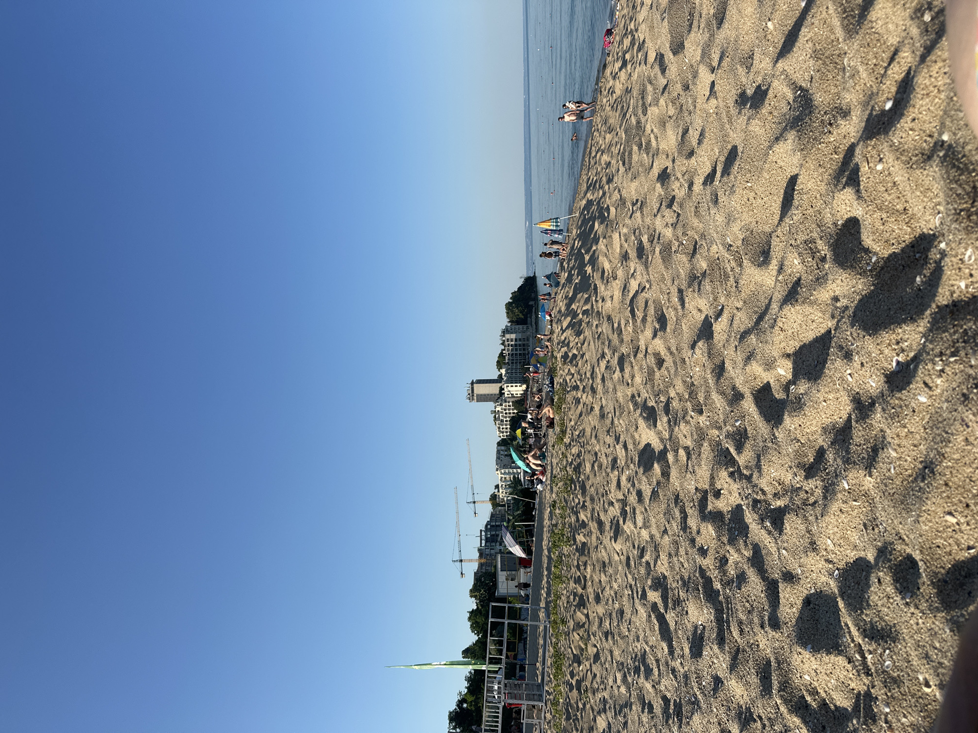 The image shows a sandy beach on a clear, sunny day. The sky is a vibrant blue with no clouds. The sand occupies the majority of the foreground, with clear footprints and shadows cast by an unseen object.
In the middle ground, a line of buildings is visible, suggesting an urban or resort area adjacent to the beach. Several construction cranes are also present, indicating ongoing development. Beach umbrellas and a few people are scattered along the shoreline. The sea stretches out to the horizon, with a few small boats visible.
The overall scene depicts a typical day at the beach, with people enjoying the sun and sea. The presence of construction suggests a potentially developing or popular tourist destination. The time of day appears to be late morning or early afternoon, given the bright sunlight and the length of the shadows. The mood is one of leisure and relaxation. There is no visible text in the image.