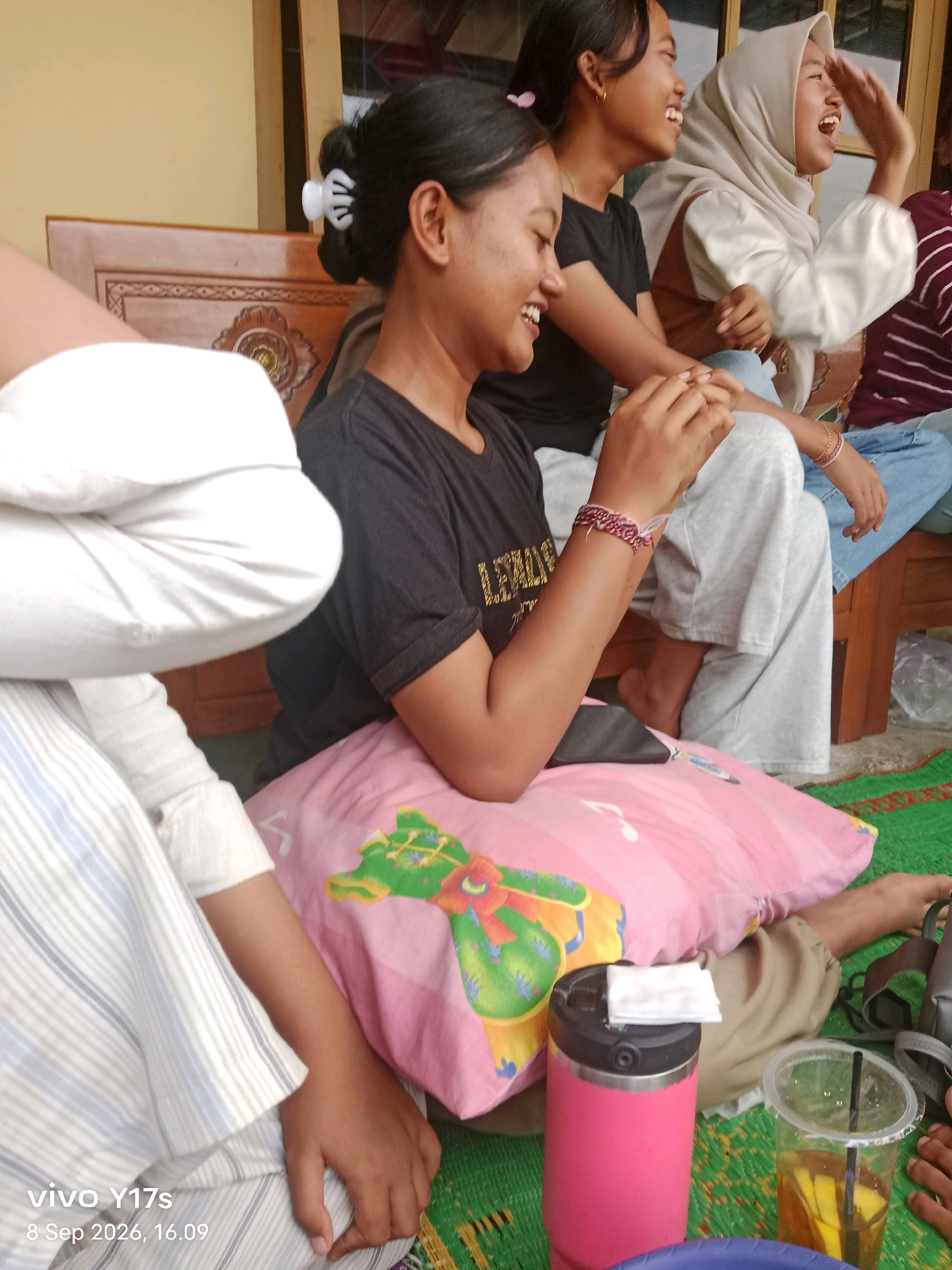 Four young women are seated on a mat outdoors, in what appears to be a residential area. The woman in the foreground on the left is wearing a black t-shirt and has a white hair clip. Next to her, a woman in a black t-shirt with gold lettering laughs. To her right, a woman wearing a light-colored hijab and a white top also laughs, her right hand raised. The fourth woman, partially visible, wears a blue shirt and is looking down. They are sitting on a mat in front of a wooden bench, with a pink pillow on the ground. A pink thermos and a clear plastic cup with a yellow beverage and straw are also present. The background includes a wooden structure and a wall. The timestamp "vivo Y17s 8 Sep 2026, 16.09" is visible in the lower left corner. The location is identified as Rama Dewa, Indonesia.