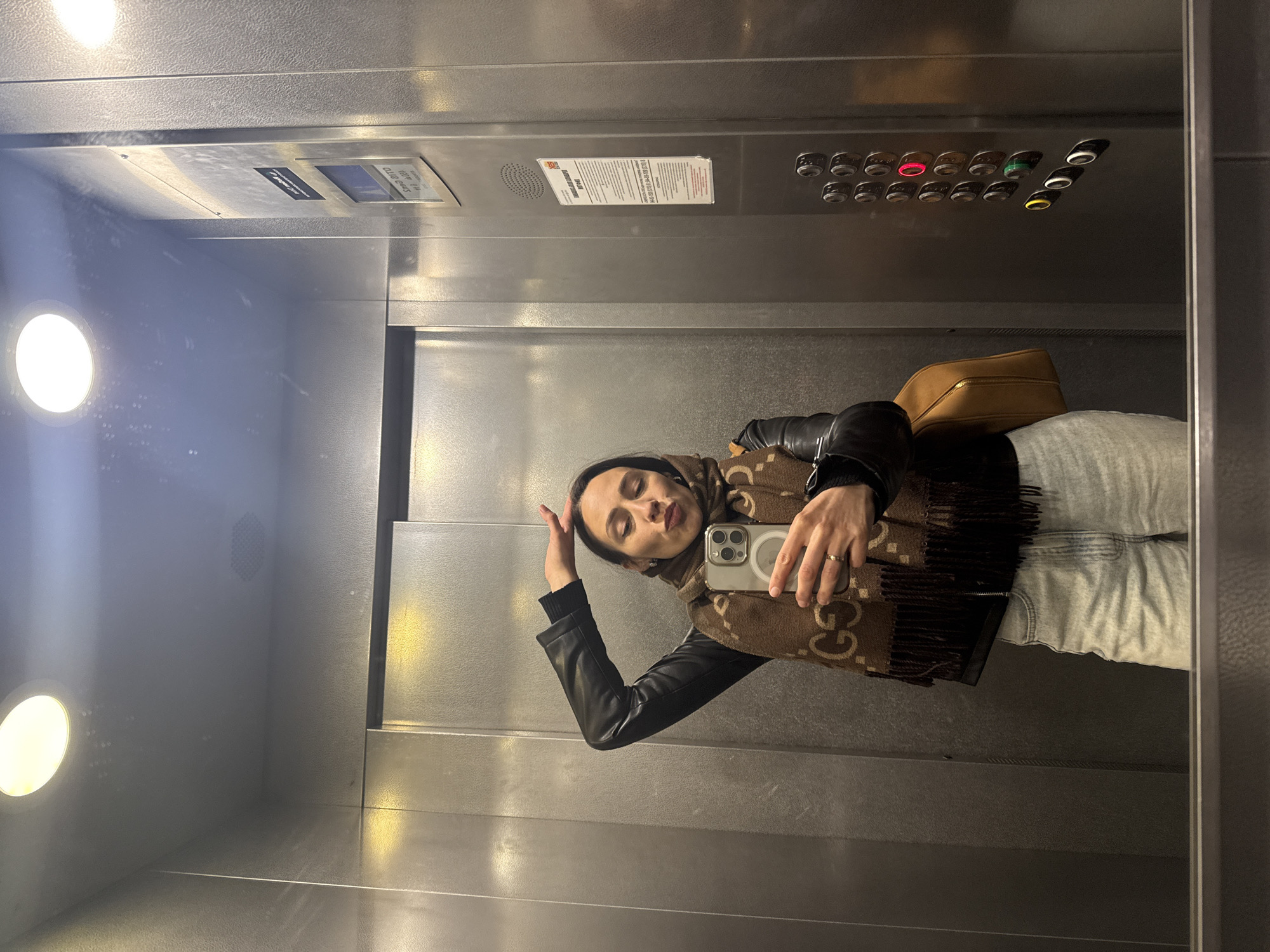 A woman is taking a selfie inside an elevator. She is wearing a black leather jacket, a brown scarf with the Gucci logo, and light-colored jeans. She is holding a smartphone in her right hand. The elevator has metal walls, a panel with floor buttons, and a light fixture. The floor buttons suggest the elevator serves many floors, with one button glowing red. The location is the city of Saint Petersburg, Russia.