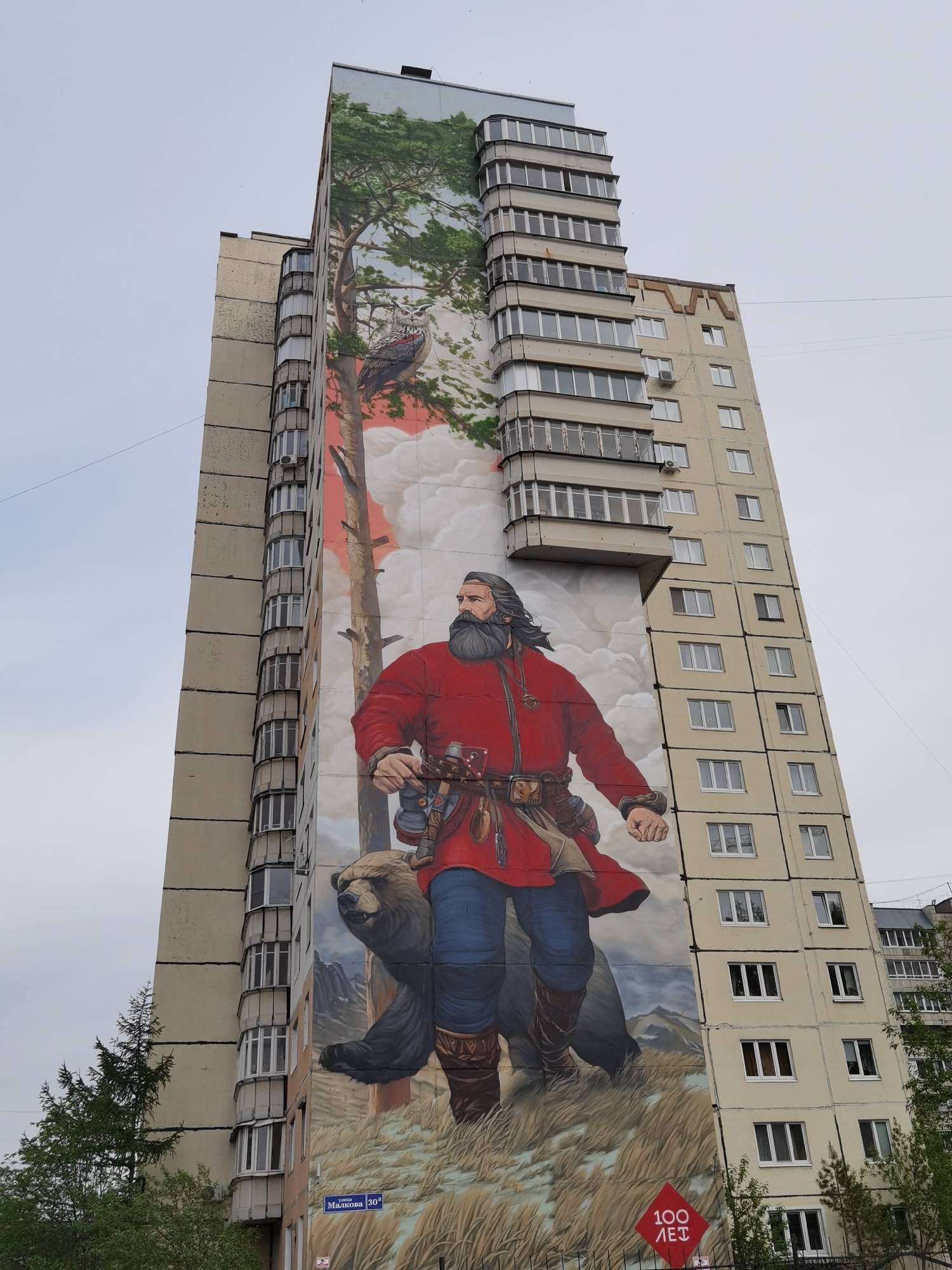 The image shows a tall apartment building in Perm, Russia, adorned with a large mural on one side. The mural depicts a bearded man in a red coat and blue pants, accompanied by a brown bear, walking through a grassy landscape with mountains in the background. A tree with an owl perched in its branches extends to the top of the building. A street sign is visible at the bottom of the building, reading "Зота Малкова 30А", indicating the location of the building. A red sign is located below the street sign with a text of 100 years. The sky is overcast.