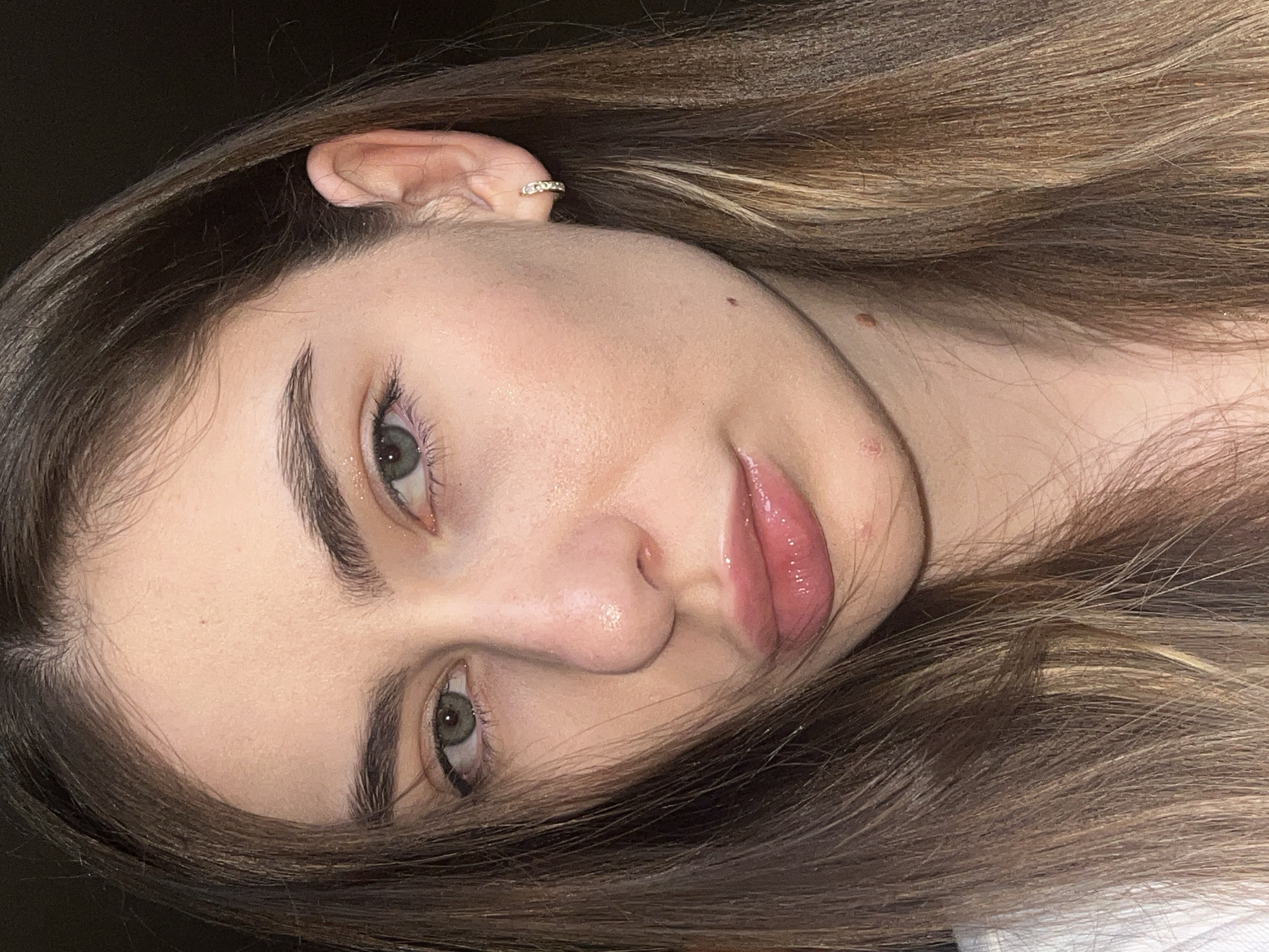 A close-up, rotated image depicts a fair-skinned individual with brown hair. The individual's eyes are green and their lips are coated with a glossy substance. A small, gold hoop earring is visible in the ear. Two small moles are on the face. The background is a dark, solid color. The location is not discernible from the image.
