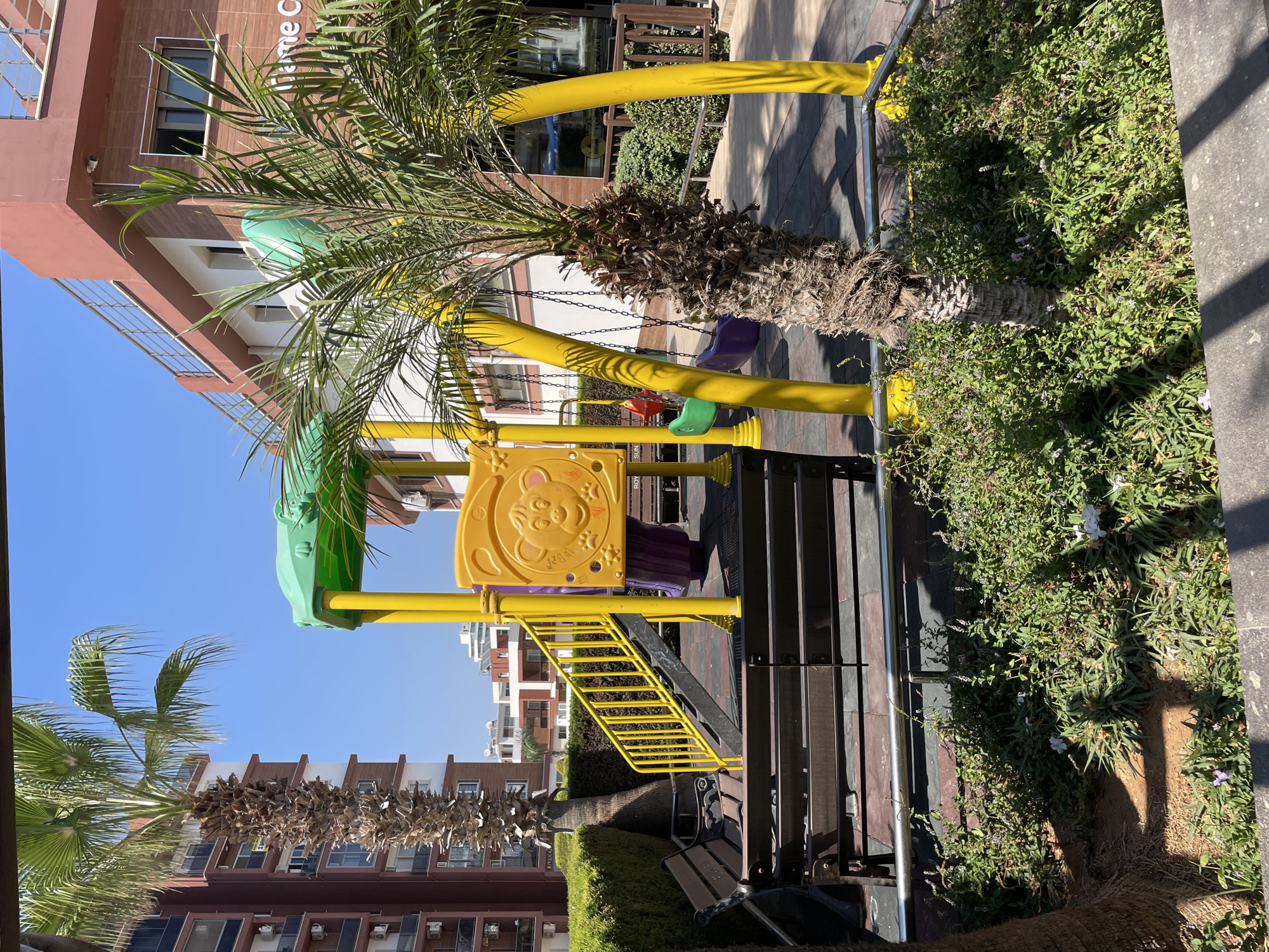 The image depicts a children's playground in front of a modern apartment building. The playground features a yellow and green plastic structure with a slide and swings. Yellow metal poles support the structure, and purple and green swings hang from chains. A yellow ladder leads up to the play structure. In the foreground, two dark wooden benches with metal legs are positioned on a checkered rubber surface. Lush green bushes and a palm tree with a textured trunk are prominent in the scene. The apartment building behind the playground is multi-story with a red-brown facade and white accents, featuring balconies and windows. The sky is clear and blue, suggesting a sunny day. The location is identified as Trikomo, Northern Cyprus / Cyprus, with building signage indicating "Home C".