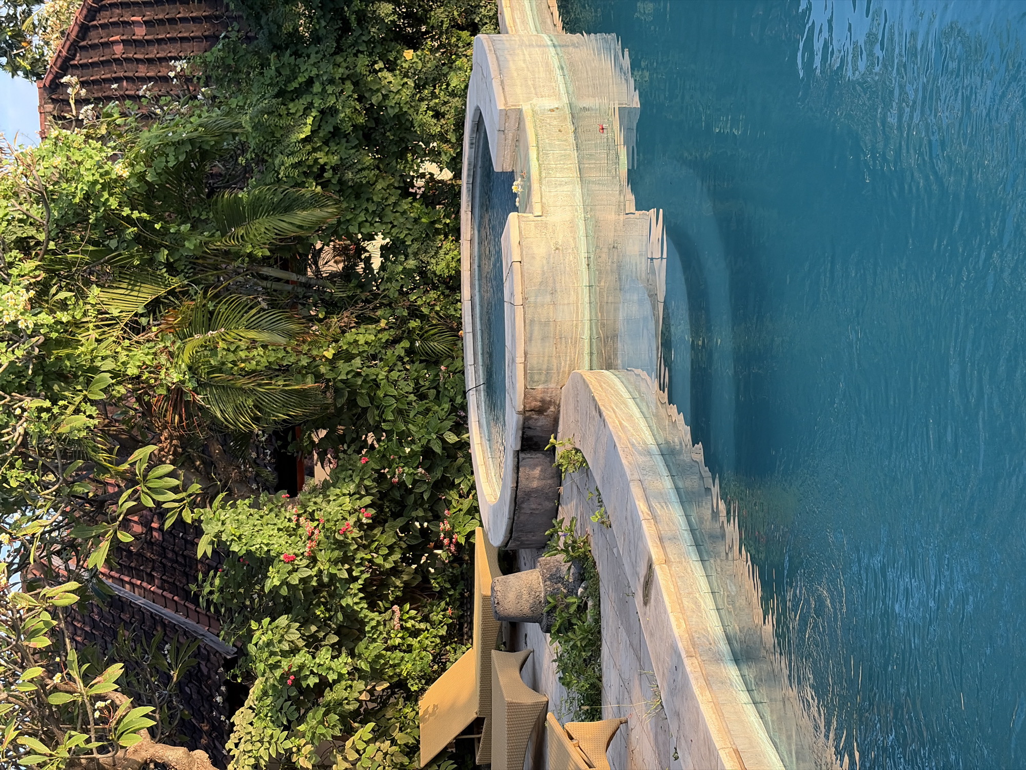 A photograph captures a resort pool area in Abang, Indonesia, bathed in the warm light of late afternoon. A bright blue swimming pool dominates the foreground, its surface rippling gently. To the left, a tan lounge chair is visible, with another out of frame. Behind the pool, a circular hot tub with a stone border is set into a tiled patio. Lush green foliage and a portion of a red-tiled roof are visible in the background. The scene is serene and tranquil.