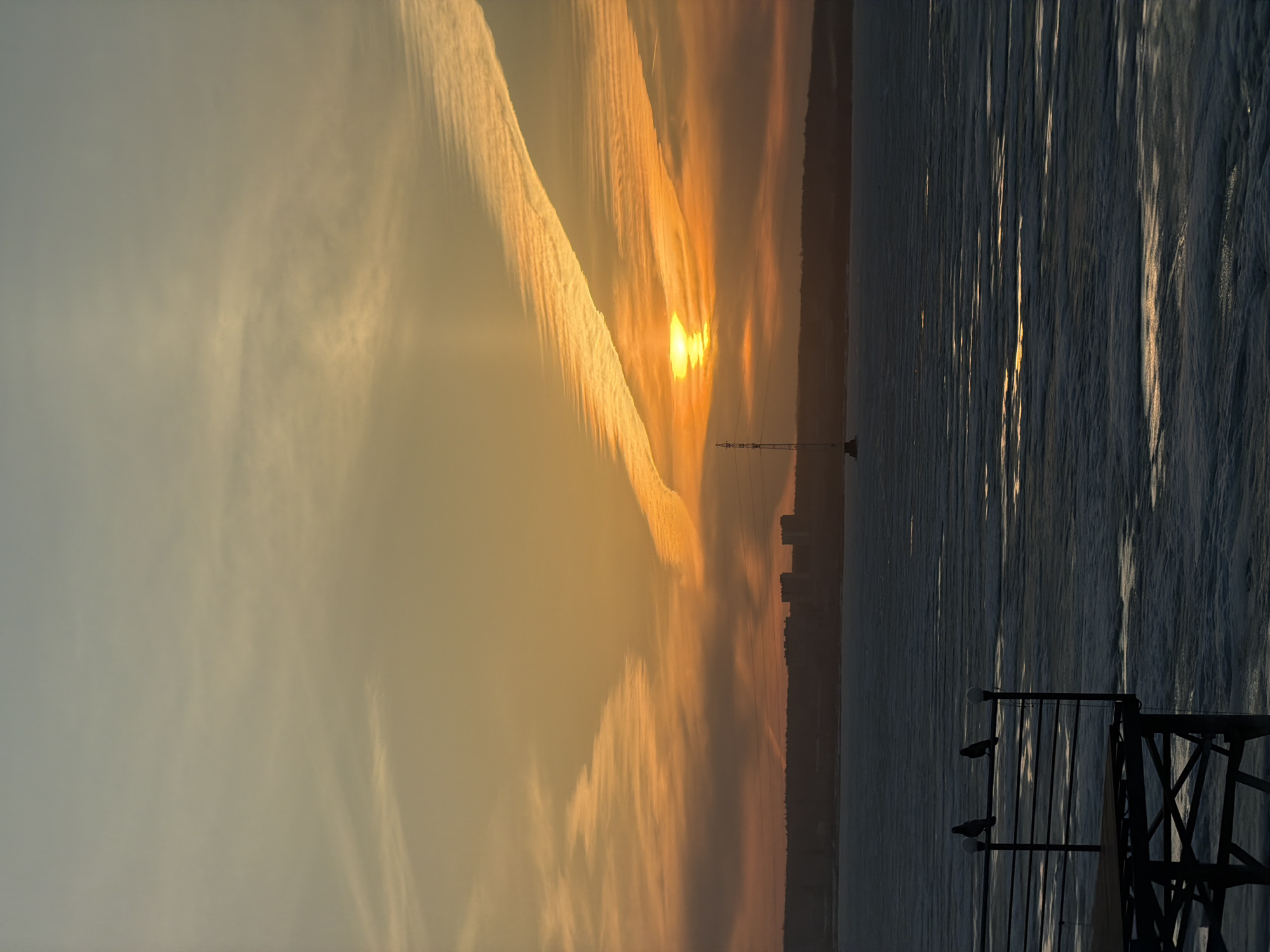 The image depicts a sunset over the Izhevsk Reservoir in Izhevsk, Russia. The sun is partially obscured by thick, striated clouds, creating a golden glow. In the distance, the silhouetted skyline of the city is visible, including an electrical transmission tower. The reservoir's surface is textured with light reflecting off the water, and in the foreground, a metal railing is present. Two birds are perched on the railing.