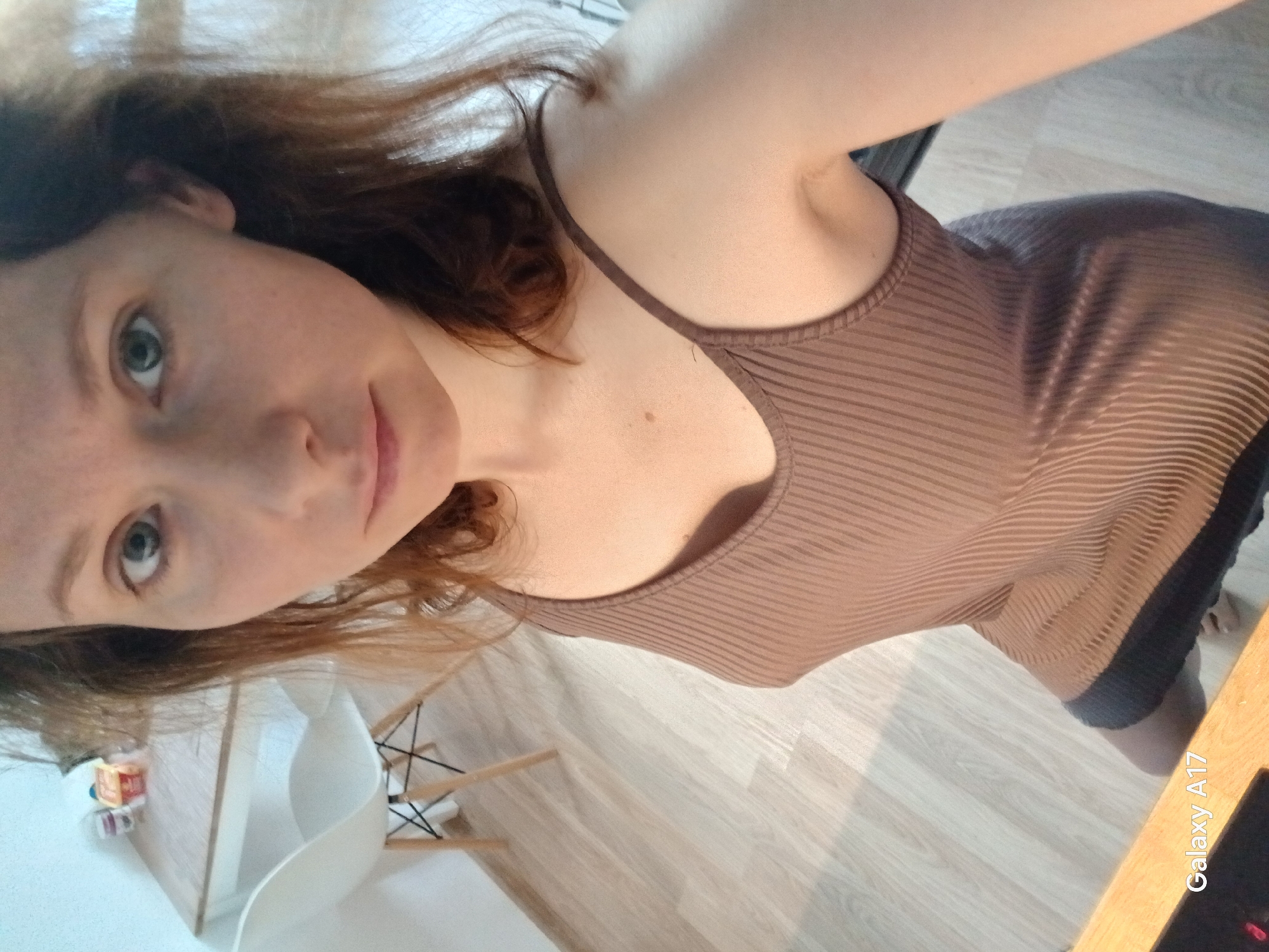 A fair-skinned woman with red hair, freckles, and green eyes is seen in a close-up selfie. She is wearing a brown ribbed tank top. She is indoors on a wooden floor. A white chair with wooden legs is visible in the background, along with some colorful items on a surface. The image also has "Galaxy A17" printed in white on the lower right corner. There is no indication of the city of Yekaterinburg or any other location in Russia.