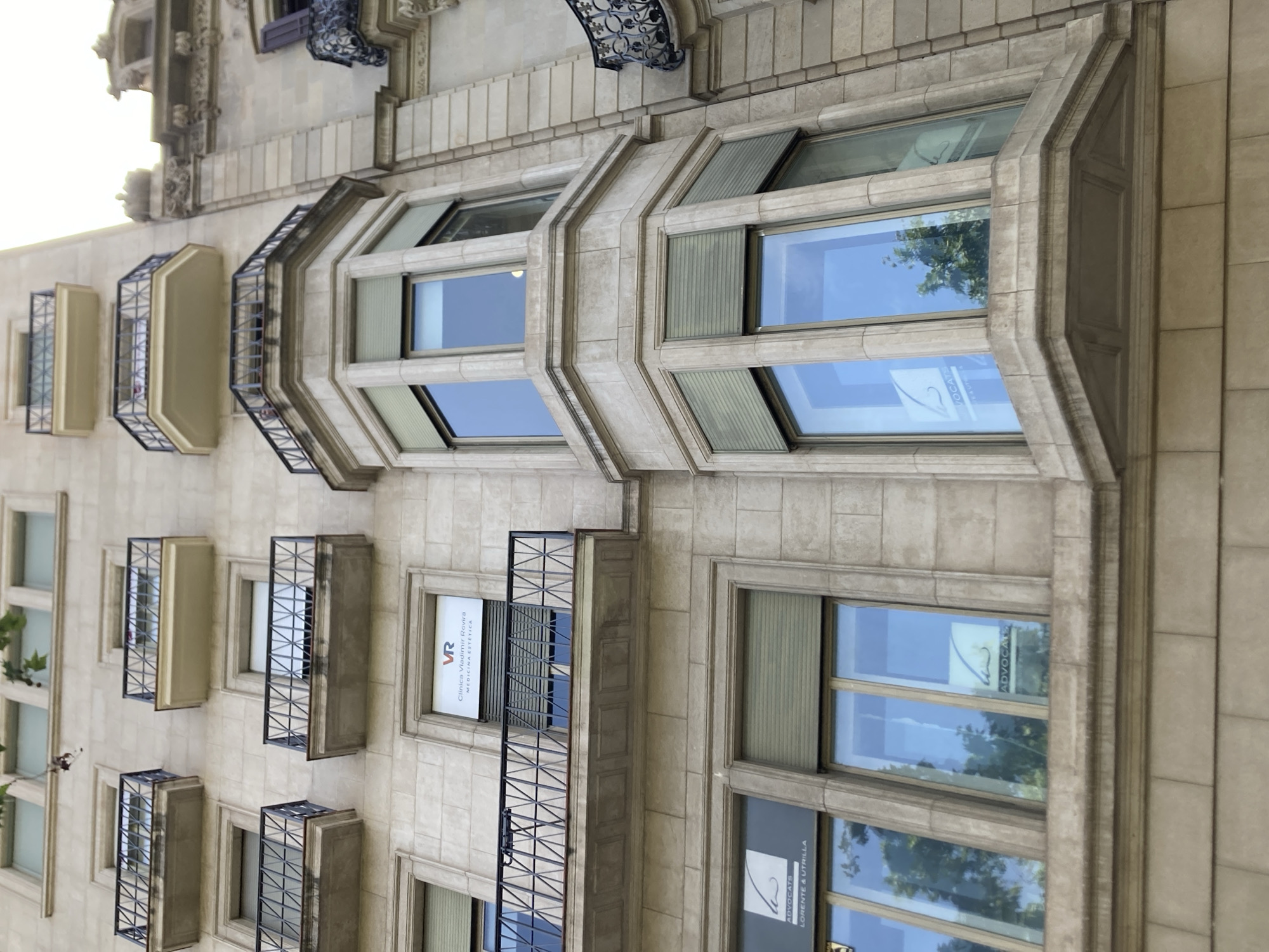 The image shows a close-up, angled view of the facade of a stone building. The architecture is ornate, with decorative elements around the windows and balconies. There are multiple windows visible, some of which are bay windows with glass panes that reflect the sky and trees. Several balconies with black metal railings project from the building. The time of day appears to be daytime, with bright sunlight illuminating the scene, suggesting clear weather.
On some of the windows, there are signs or decals with text. One window displays "VR CLINICA VLADIMIR-ROVIRA MEDICINA ESTÉTICA". Another window shows "ADVOCATS LORENTE & UTRILLA". There are also other, less legible signs or logos within the windows.
The overall impression is of a well-maintained, possibly historic building in an urban setting, likely in Barcelona, Spain, given the architectural style and the presence of Spanish text on the signs. There are no people visible in the image.