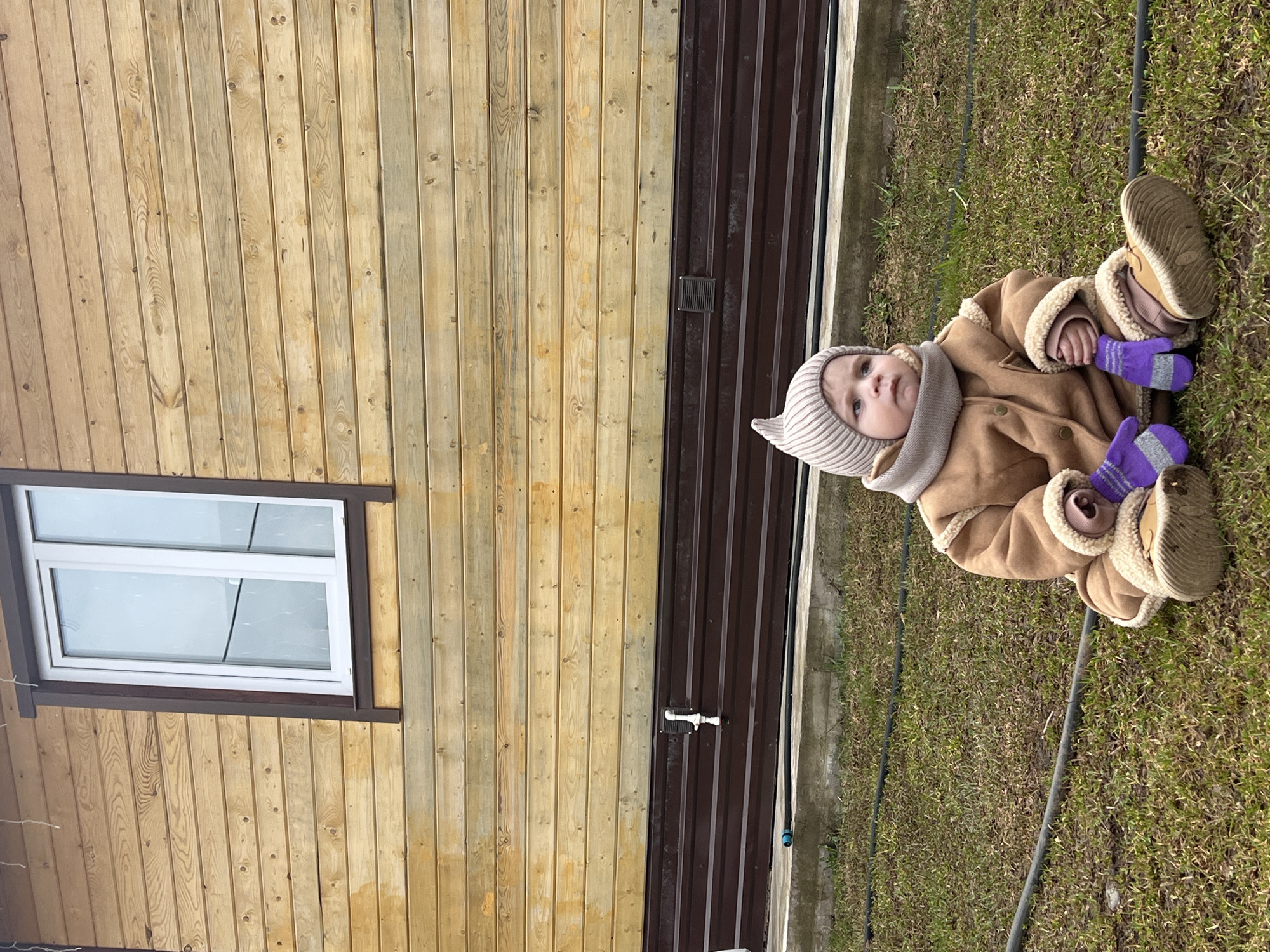 A young child, appearing to be a toddler, is seated on green grass in an outdoor setting, likely a backyard in Protasy, Russia. The child is warmly dressed for cool weather, wearing a light-colored ribbed beanie, a tan shearling-lined coat with lighter trim, a coordinating scarf, and striking dark purple mittens with gray stripes. Their footwear consists of sturdy light brown boots. The child's gaze is directed slightly upwards and to the left, showing a calm and curious expression.
The background features a structure composed of vertical wooden planks, possibly a house or shed, with a white-framed window visible on its left side. To the right of the wooden structure, a dark brown corrugated metal gate or wall extends vertically, featuring a small square handle or latch. The ground is predominantly green grass, with some drier patches, and a black hose snakes across the lower part of the scene. The lighting suggests it is daytime, with clear, mild weather. No text is visible in the image.