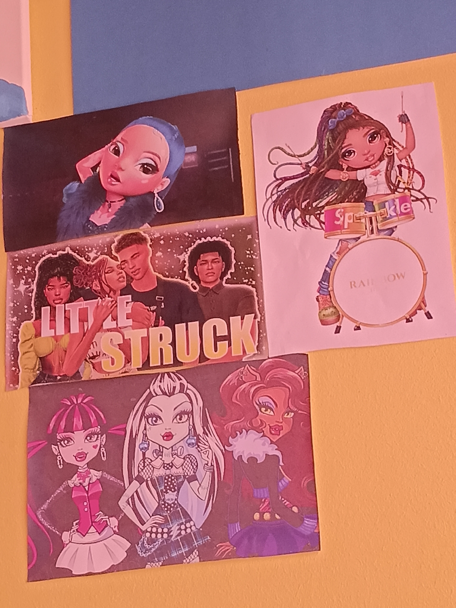 The image displays a collection of four stickers or prints adhered to a wall. From top to bottom and left to right, the stickers are:
1. A close-up of a doll-like character with blue hair, large eyes, and wearing a blue fur collar.
2. A sticker of four people. Two are in the foreground embracing, with the text "LITTLE STRUCK" prominently displayed. Two more figures are visible behind them.
3. A stylized illustration of a doll with long, dark, wavy hair, playing drums. The drums have the word "Sparkle" on them, and the larger drum has "RAINBOW" written on it.
4. An illustration featuring three doll-like characters, seemingly from the Monster High franchise. The characters are depicted with distinct hairstyles and outfits: one with pink hair in pigtails, another with long white and black hair, and a third with long red hair.
The stickers are arranged on a wall that appears to be painted in two sections: a yellow-orange lower section and a blue upper section. The image is a close-up shot, and the stickers are the primary focus. There is no visible information or context within the image that would indicate its location in Wels, Austria.