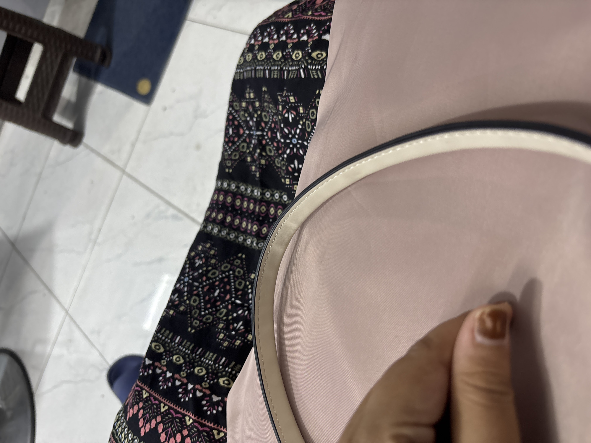 In Wanajaya, Indonesia, a close-up shot shows a person's hand resting on a pale pink fabric, possibly a dress or a bag. The hand has short, unpolished fingernails. A thin, off-white strap with dark edging, likely from a handbag, curves across the pink material. In the background, a black fabric with a colorful geometric pattern is visible, along with a portion of a blue flip-flop. The floor is tiled in a light, neutral color. A dark brown stool is partially visible in the upper right corner, with a dark blue mat beneath it.