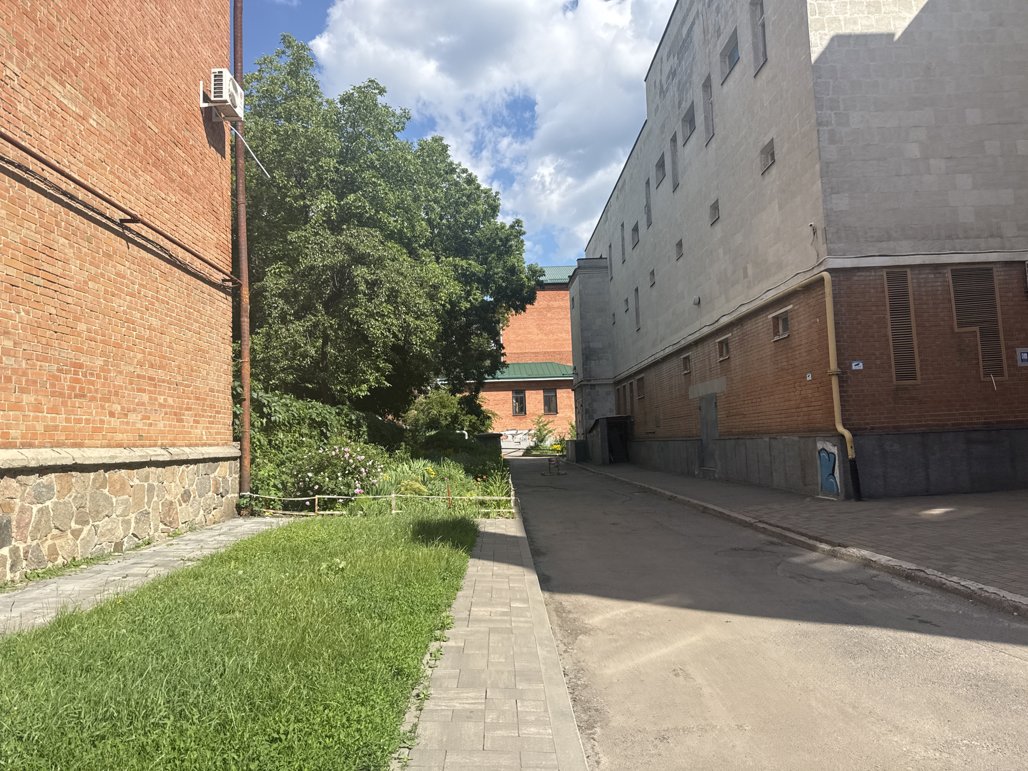 The image depicts a narrow alleyway between buildings, oriented vertically. To the left, a paved walkway runs alongside a well-maintained grassy area. Beyond the grass, a rough-hewn stone wall borders a red brick building. Further into the background, a large deciduous tree with dense green foliage dominates the scene, with some pink and yellow flowers visible beneath it. Adjacent to the tree is another red brick building. To the right of the alley, a tall, light-colored building with numerous square windows rises, its facade appearing to be made of a concrete or stucco material. A brown pipe runs along the side of the building. On the top of the brick building, an air conditioning unit is mounted. The sky above is a clear blue with scattered white clouds. The lighting suggests it is daytime with the sun casting shadows.