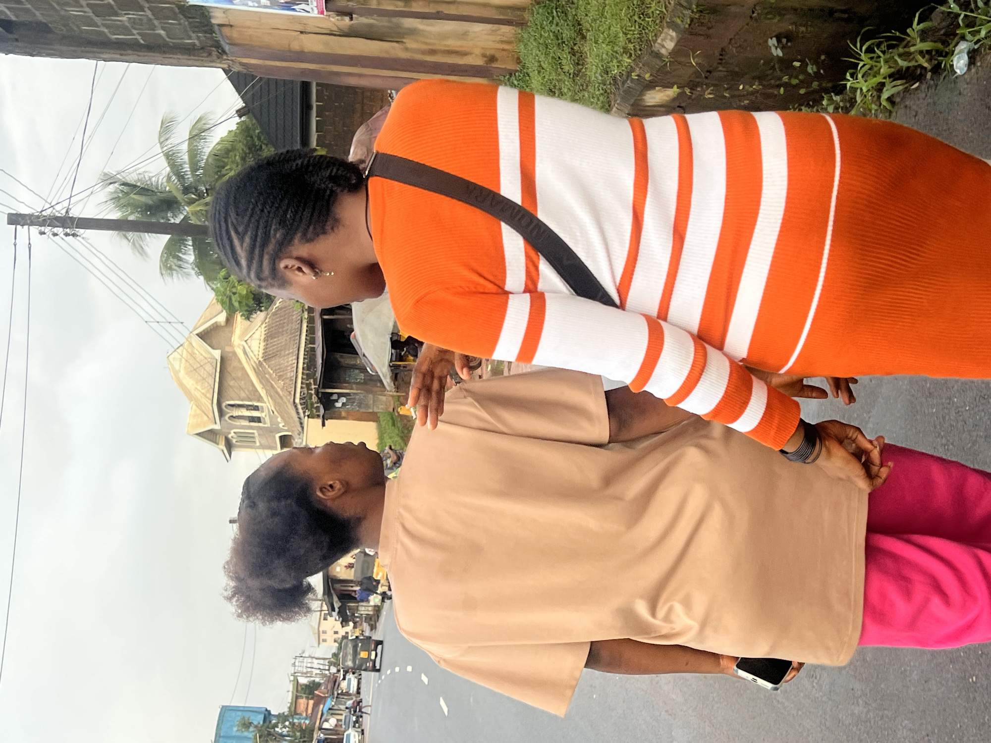 Two women are walking on a paved road in Nkpor, Nigeria. The woman on the left is wearing a tan t-shirt and bright pink pants. She is holding a smartphone in her left hand. The woman on the right is wearing a long-sleeved orange and white striped dress and a black crossbody bag. She has braided hair and is wearing small hoop earrings. The women are walking away from the camera and appear to be conversing. The background shows buildings with corrugated metal roofs, a palm tree, power lines, and a few vehicles on the road. The sky is overcast. The scene depicts a street in an urban setting.