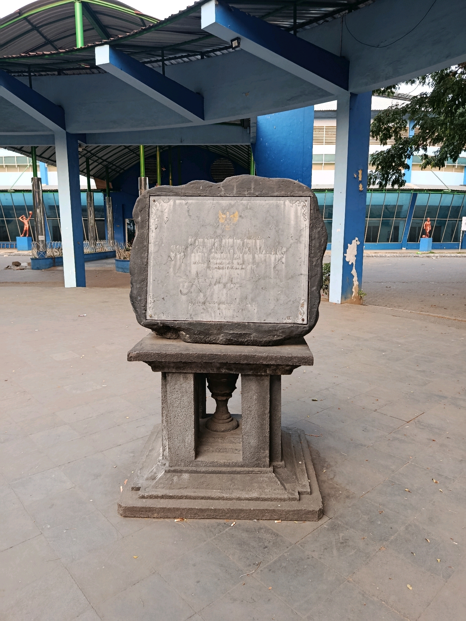 This image depicts a monument located in front of the GOR Ken Arok in the city of Malang, Indonesia. The monument consists of a large, rough-hewn stone slab with an inscribed plaque, mounted on a tiered stone pedestal. The plaque features a national emblem and Indonesian text. The surrounding environment includes a paved area with stone tiles, a covered structure with blue and white columns and a metal roof, and a building with blue glass windows in the background. There are also some decorative statues of athletes visible in the background, as well as trees and foliage. The scene appears to be outdoors during the daytime, with bright lighting.