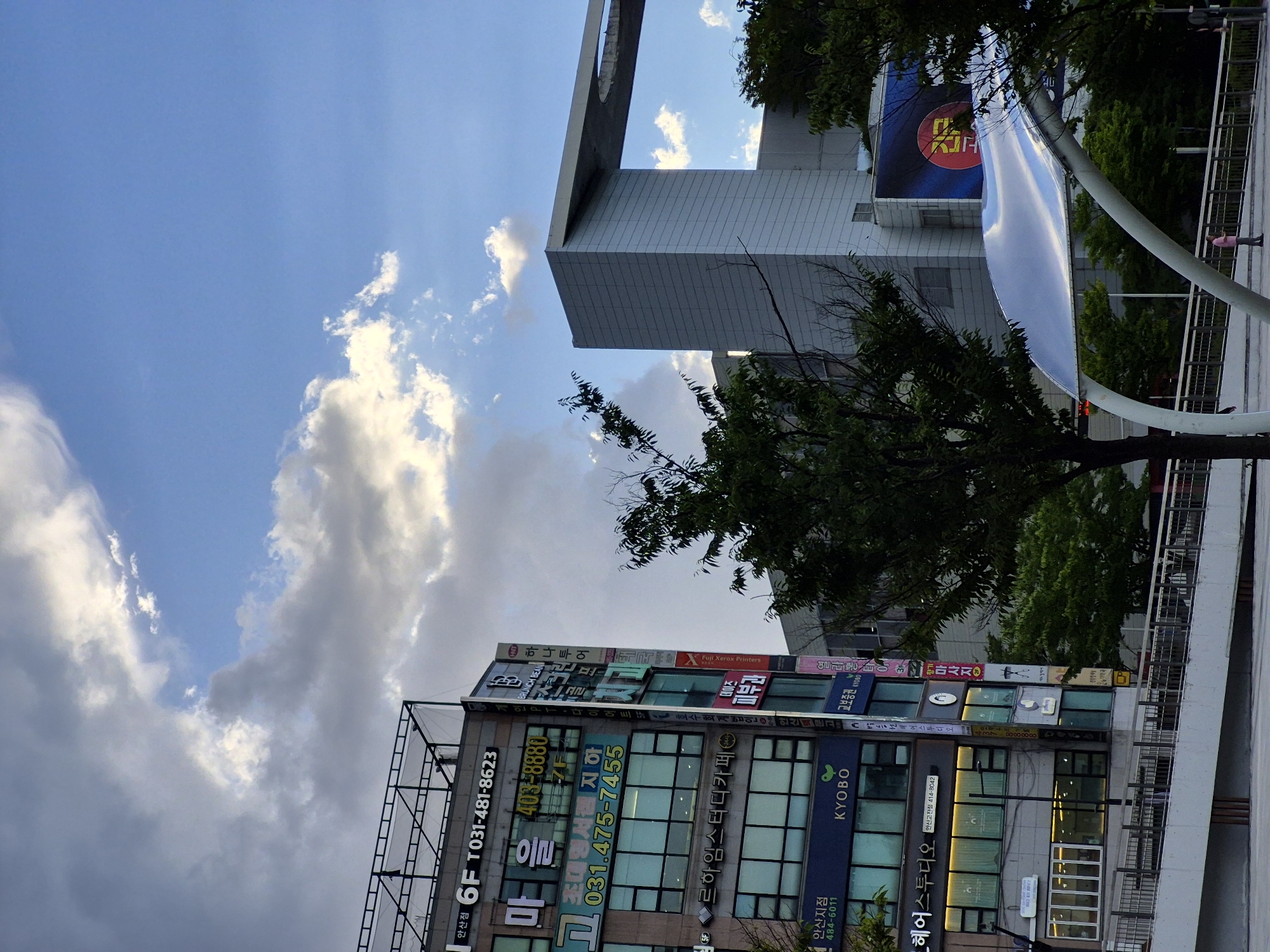 The image shows a street scene in Ansan, South Korea, with a focus on buildings and the sky. In the foreground, there is a white, modern-looking pedestrian ramp with a metal railing. Beyond the ramp, several multi-story buildings are visible. The building on the left is clad in gray panels and features numerous signs with Korean text and numbers, indicating businesses such as a hair salon ("헤어스튜디오") and a bookstore ("KYOBO"). The building on the right is a taller, modern structure with a gray facade and a large, arched overhang. The sky above is a mix of blue sky and scattered clouds, with some clouds appearing bright and illuminated by the sun. Trees with green foliage are interspersed throughout the scene, partially obscuring some of the buildings. A single person wearing a pink shirt and dark pants is visible standing on the pedestrian ramp in the middle ground. The overall atmosphere suggests an urban environment on a partly cloudy day.
