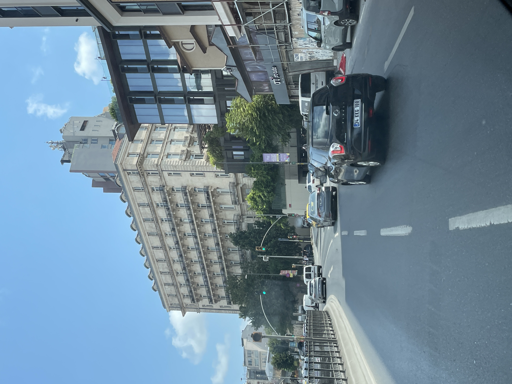 The image depicts a street scene in Beyoğlu, Turkey, on a clear day with scattered clouds. In the foreground, a black SsangYong Tivoli SUV with the license plate "34 KKG 188" is driving on a multi-lane road. Other vehicles, including a silver car and vans, are visible in the traffic. The road is lined with a metal fence on the left and various buildings on the right. Behind the fence, a tram line is present. The buildings in the background are a mix of architectural styles, including a large, ornate stone building with numerous balconies and arched windows, and a modern glass-fronted building. Trees and traffic lights are also part of the urban landscape. The overall impression is that of a busy city street with active vehicular and pedestrian traffic.