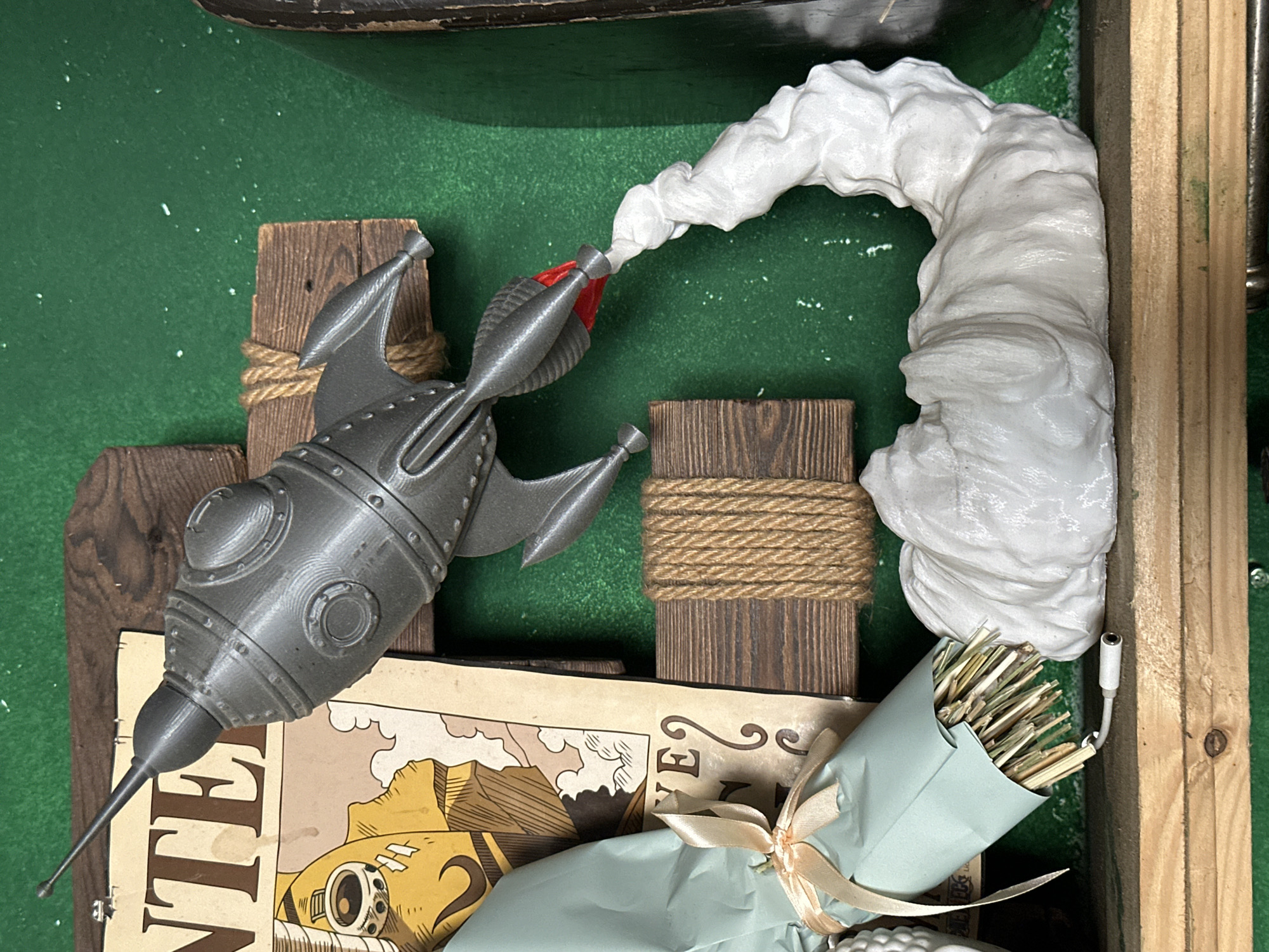 The image shows a diorama of a retro-style rocket ship taking off. The rocket is metallic gray with a red accent at the nozzle and appears to be 3D printed. It rests on top of a vintage-looking map or poster. The rocket is positioned on two small wooden posts, with twine wrapped around them. A cloud of white material simulates the rocket's exhaust. To the right of the cloud is another small wooden post wrapped with twine, and next to it is a bouquet of dried grass or straw wrapped in light blue paper and tied with a ribbon. The background is a solid green surface, and portions of wooden framework are visible around the edges of the image. The location, Andreyevka, Russia, is not determinable from the visual information.