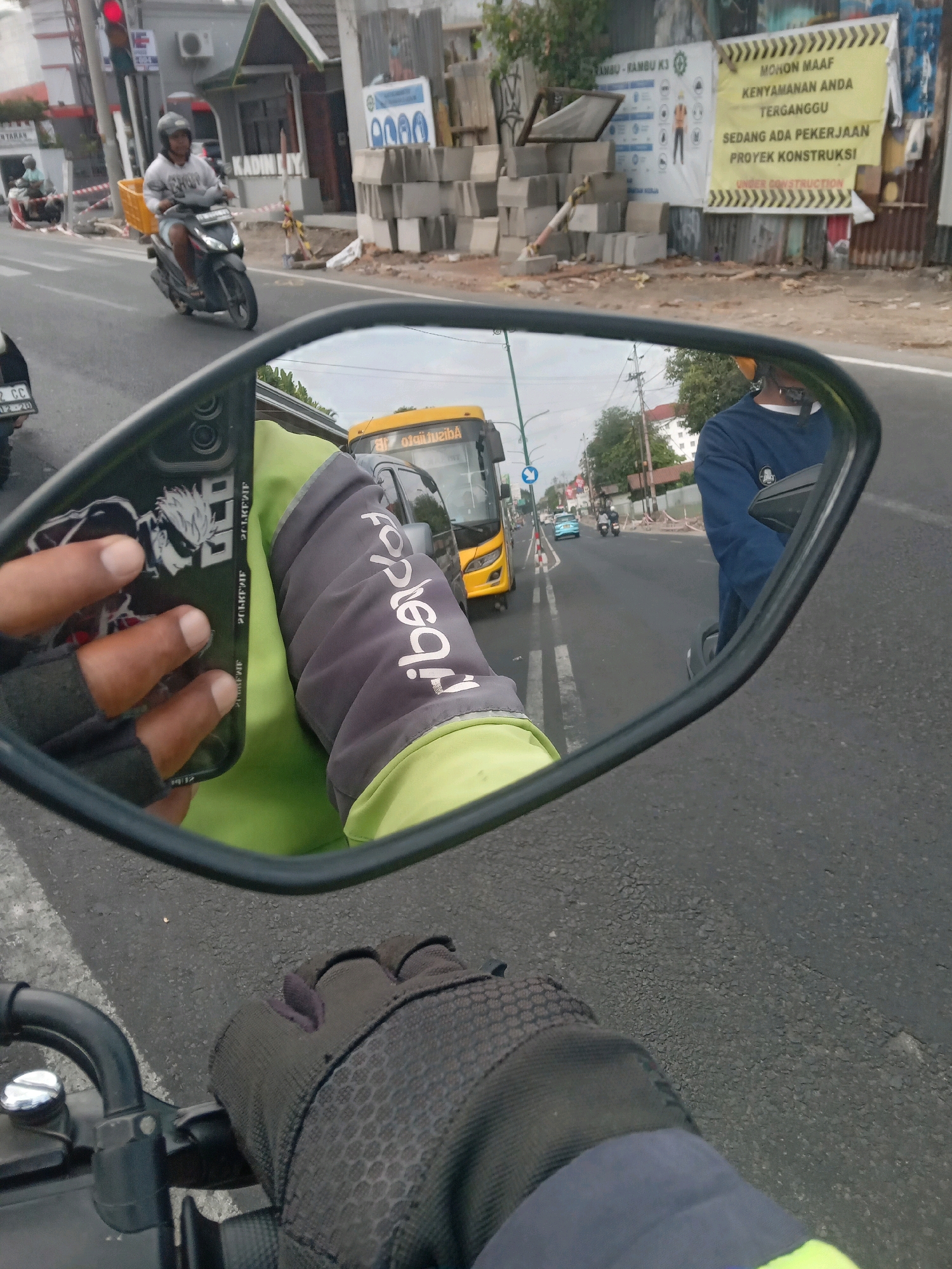 The image captures a first-person perspective from a motorcycle in Yogyakarta, Indonesia, looking into the side mirror. The mirror reflects a busy street scene with traffic. In the foreground, a hand wearing a grey glove is visible, holding a smartphone with a dark cover featuring an anime character. The rider is wearing a bright green and grey jacket.
Reflected in the mirror, a yellow bus is prominent, driving on the road. Other vehicles, including a blue car and two motorcycles, are also present in the traffic. On the side of the road, construction materials, including stacks of gray bricks, are visible, along with construction signage. One sign, in yellow, reads "MOHON MAAF KENYAMANAN ANDA TERGANGGU SEDANG ADA PEKERJAAN PROYEK KONSTRUKSI" which translates to "We apologize for the inconvenience, construction work is in progress." This suggests the location is near an ongoing construction project. In the background, buildings and trees are seen, indicating an urban environment. A traffic light is also visible on the left side of the image.