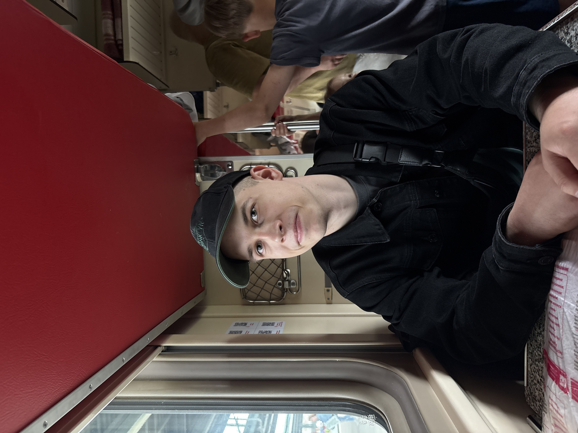 A young man wearing a black baseball cap and black denim jacket is seated inside a train compartment, looking towards the camera. He has a slight smile and is resting his arms on a table covered with a patterned sheet. The compartment has beige walls, a red ceiling, and a window to his left showing a platform with people and buildings outside. Other passengers, including a young boy in a grey t-shirt, are visible in the background. The image appears to be taken in Russia, likely on a long-distance train.
