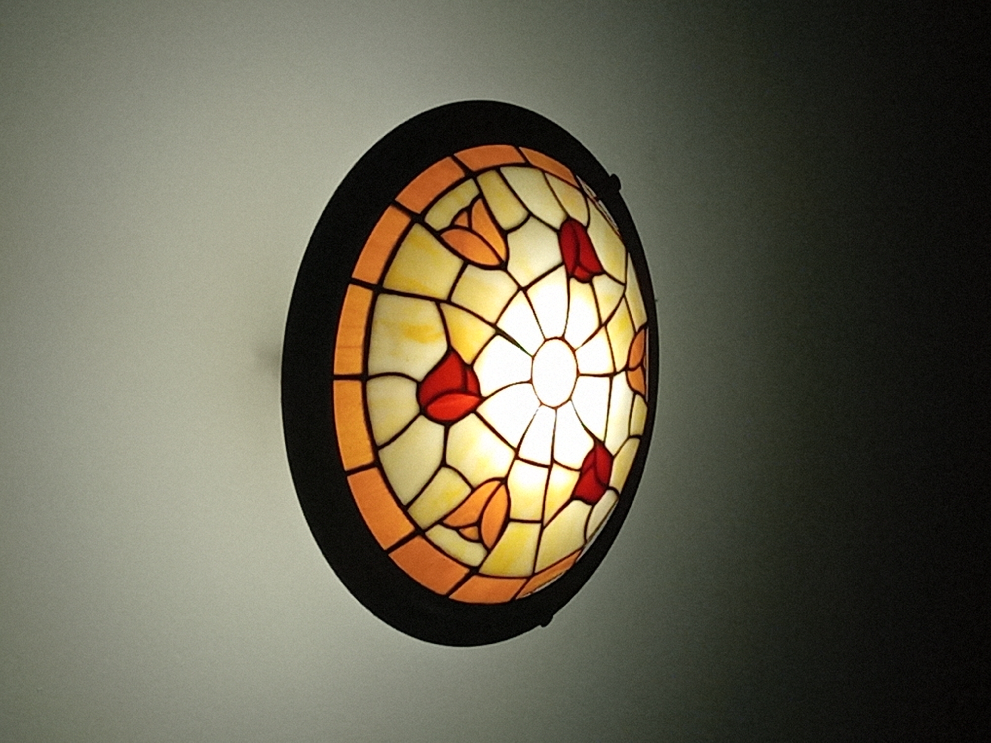 A lit, circular stained glass wall sconce with a dark frame is mounted on a light gray wall. The sconce is composed of numerous small pieces of stained glass in white, orange, and red hues. The white glass is in the center with red and orange accents. The light source is situated at the center of the fixture, casting a bright glow. The background is a gradient from light gray to dark, with the sconce positioned towards the left side of the frame. Location is unknown.