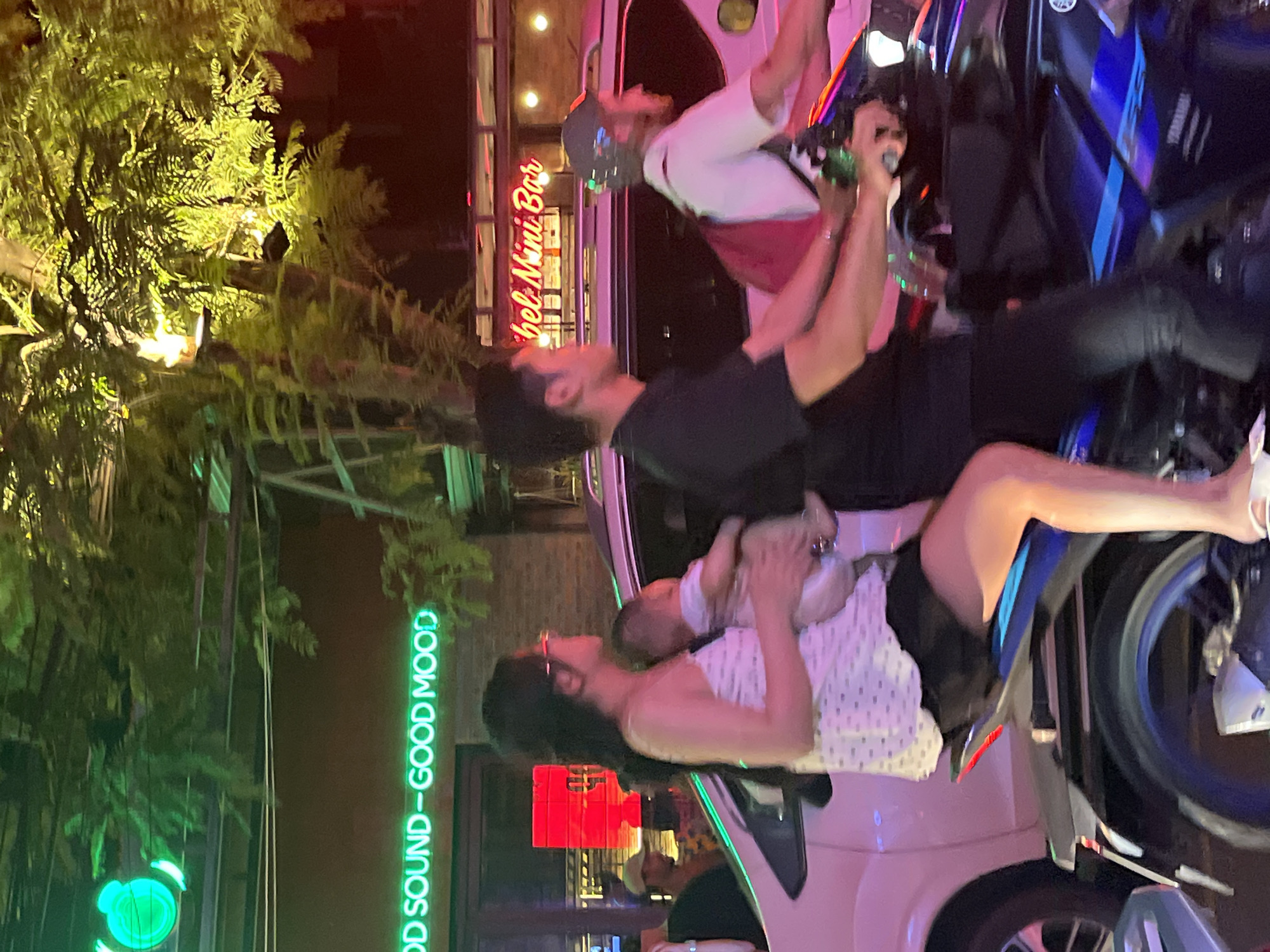 At night, a man and a woman are seated on a white car and a blue motorbike parked in front of a bar in Đà Nẵng, Vietnam. The woman, wearing a white dress with black polka dots, holds a baby. The man, wearing a black shirt, is seated behind her. Another man, wearing a white shirt with a red and black design and a baseball cap, stands near the motorbike. The bar's neon sign reads "Bel Mini Bar." Other neon signs nearby read "GOOD SOUND - GOOD MOOD." There are trees in the vicinity.