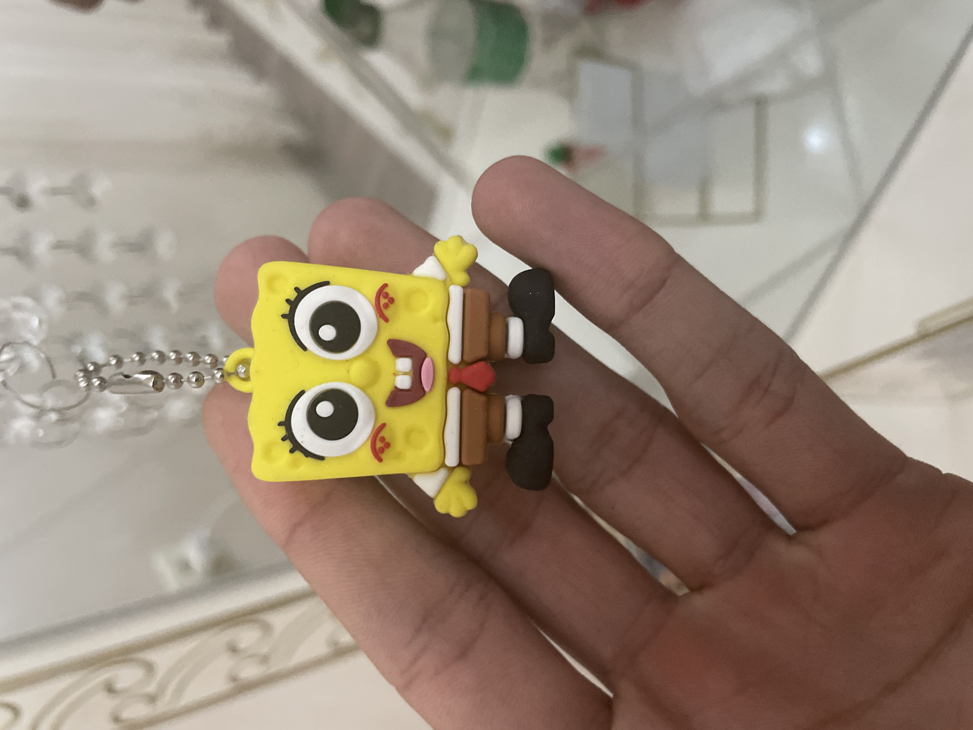 A human hand with light to medium skin tone holds a Spongebob Squarepants keychain or charm prominently in the center of the frame. The yellow, square-shaped charm depicts Spongebob with large, cartoonish eyes, an open mouth revealing two teeth and a pink tongue, and red blush marks on its cheeks. The character is outfitted with a white shirt, brown pants, a red tie, and black shoes. A silver-colored ball chain extends from the top of the charm, connected to a partially visible metal keyring. The background is out of focus, revealing a light-colored wall or furniture with ornate white detailing in the upper left. Above the charm, blurred, reflective, crystal-like objects are visible, consistent with a chandelier or decorative light fixture. To the right, an out-of-focus object with a green element, possibly a bottle, rests on a light-colored surface. The lower background suggests a light-colored, possibly tiled or polished floor. The scene is indoors and well-lit. The image contains no visual information to determine its location as Ihtiman, Bulgaria, or any other specific geographical area.