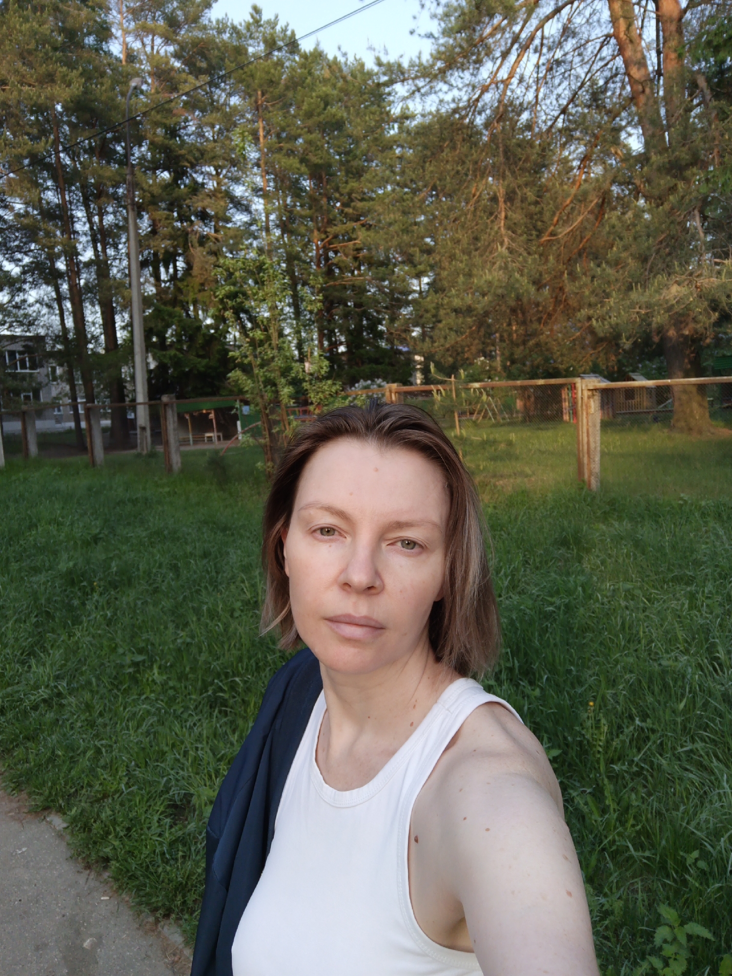Here is a detailed description of the image:
The image is a selfie taken by a woman in an outdoor setting, possibly a park or recreation area. The woman has fair skin, short brown hair, and is wearing a white tank top and a dark blue jacket draped over her shoulder. Her expression is neutral.
The background features tall trees, primarily pine trees, suggesting a wooded area. There is a fenced-in playground or open area in the middle ground with green grass. In the distance, there's a glimpse of a building, possibly a residential or public building.
Based on the prompt mentioning Chaikovsky, Russia, it is likely that this photo was taken somewhere within the city, possibly a local park or green space. The scene suggests a quiet, possibly residential area, with natural green spaces. There is no action taking place in the image. The setting is tranquil with lush green grass and forest trees. The trees are lit by late-afternoon or early-evening sunlight.