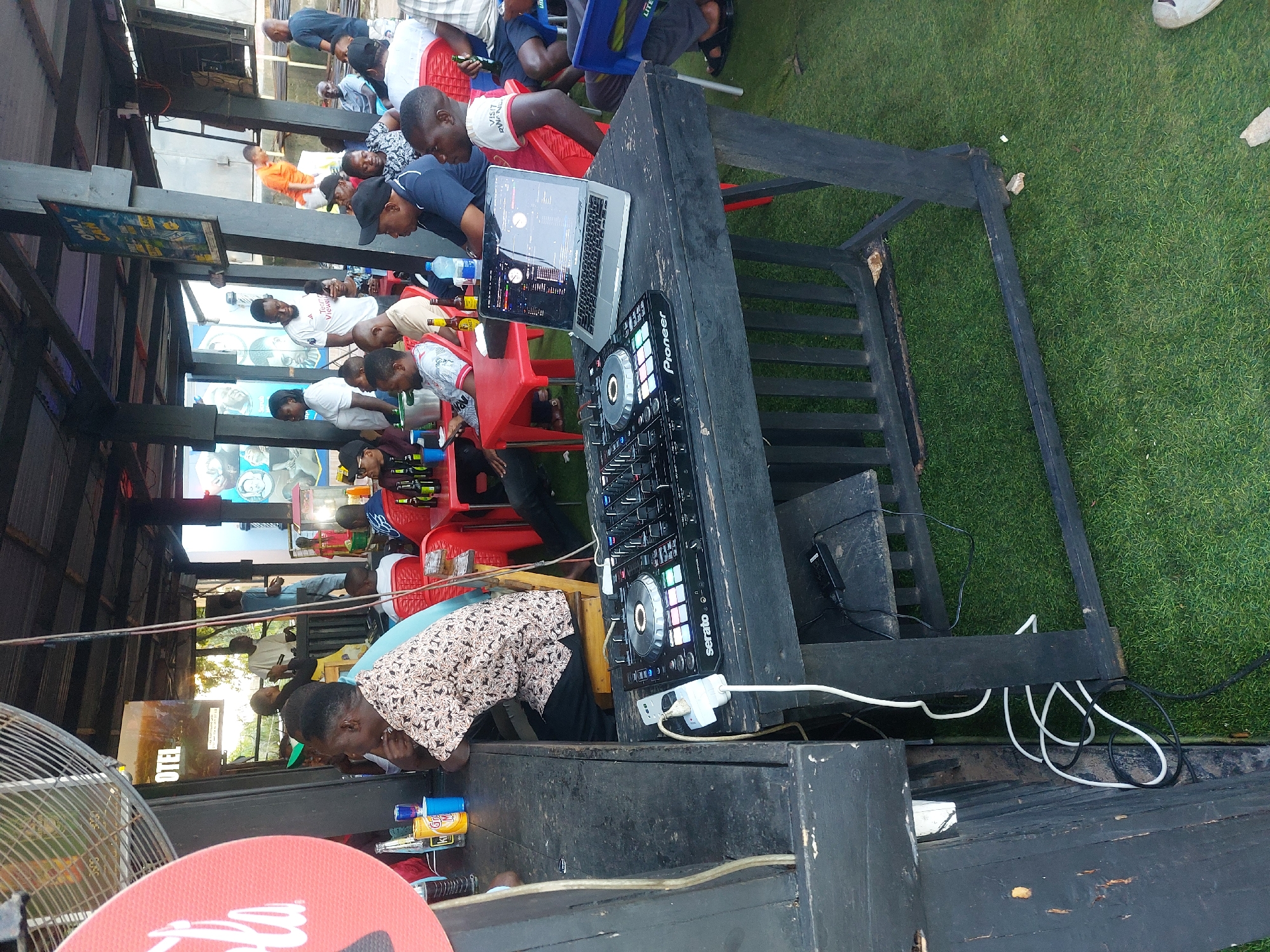 The image shows an outdoor gathering, likely in Dar es-Salaam, Tanzania. The environment includes a roofed area with supporting beams, possibly a bar or open-air restaurant, adjacent to a patch of artificial green turf. People are seated on red plastic chairs, drinking bottled beverages.
At the forefront of the scene is a DJ setup consisting of a Pioneer DJ controller and a laptop, both placed on a black wooden table. The table is positioned in front of a speaker and has electrical wires snaking around its legs. One person is close to the DJ equipment, possibly operating it. A large red advertisement sign is visible in the lower left corner, along with a standing fan.
