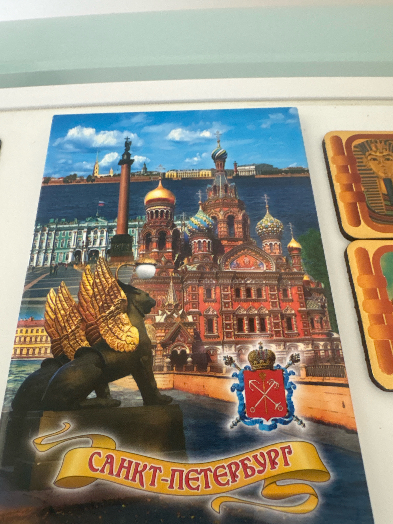 The image is a close-up, eye-level, slightly angled shot of a magnet featuring a painting of a cityscape. The painting depicts a large, ornate red building with multiple domes and spires, characteristic of Russian Orthodox architecture, situated on a body of water. To the left of this building is a grand, pale green building with classical columns and arched windows. In the foreground, a dark sculpture of a lion with large, golden wings is prominently displayed on a stone pedestal. Behind the lion sculpture is a large red and blue coat of arms. A yellow banner with Cyrillic text, reading "САНКТ-ПЕТЕРБУРГ" (Saint Petersburg), curves across the bottom of the magnet. The background shows a body of water, a distant skyline with buildings and a tall column topped with a statue. The magnet is attached to a white surface, and two other magnets, one featuring a pharaoh's head and another with a green pattern, are partially visible to the right. The location in the city of Dubovoye, Russia is not depicted or suggested in this image.