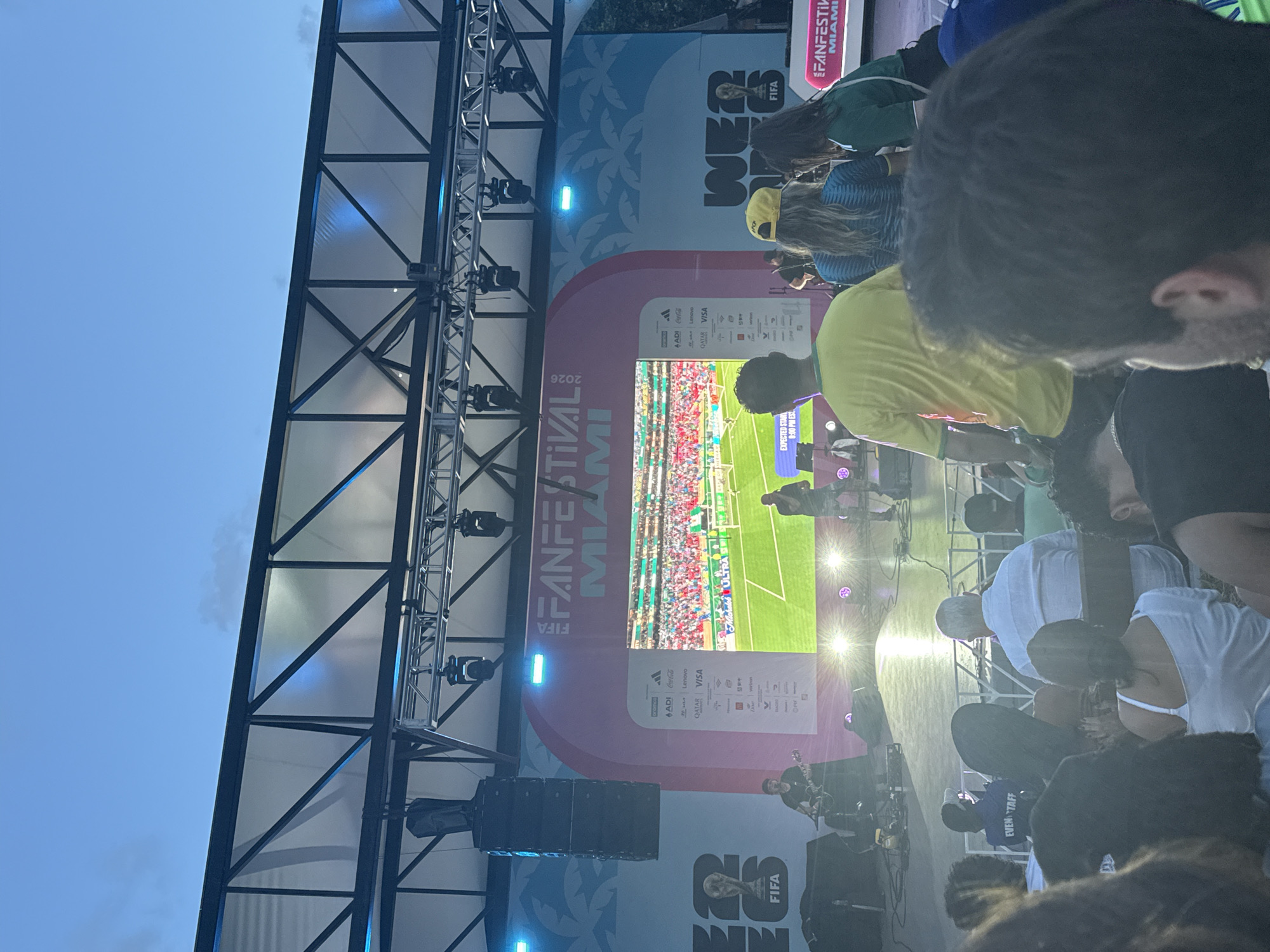 The image captures a scene at a FIFA Fan Festival in Miami, United States of America. A large screen displays a soccer match with a crowd in the stadium. In front of the screen, a stage is set up with a musician playing a guitar. Several people are gathered in front of the stage, some watching the match on the screen and others looking towards the stage. The environment appears to be an outdoor venue with a clear sky, illuminated by stage lights. The crowd includes individuals of various ages and ethnicities. The overall atmosphere suggests a lively event, likely a public viewing of a soccer game with accompanying entertainment.
